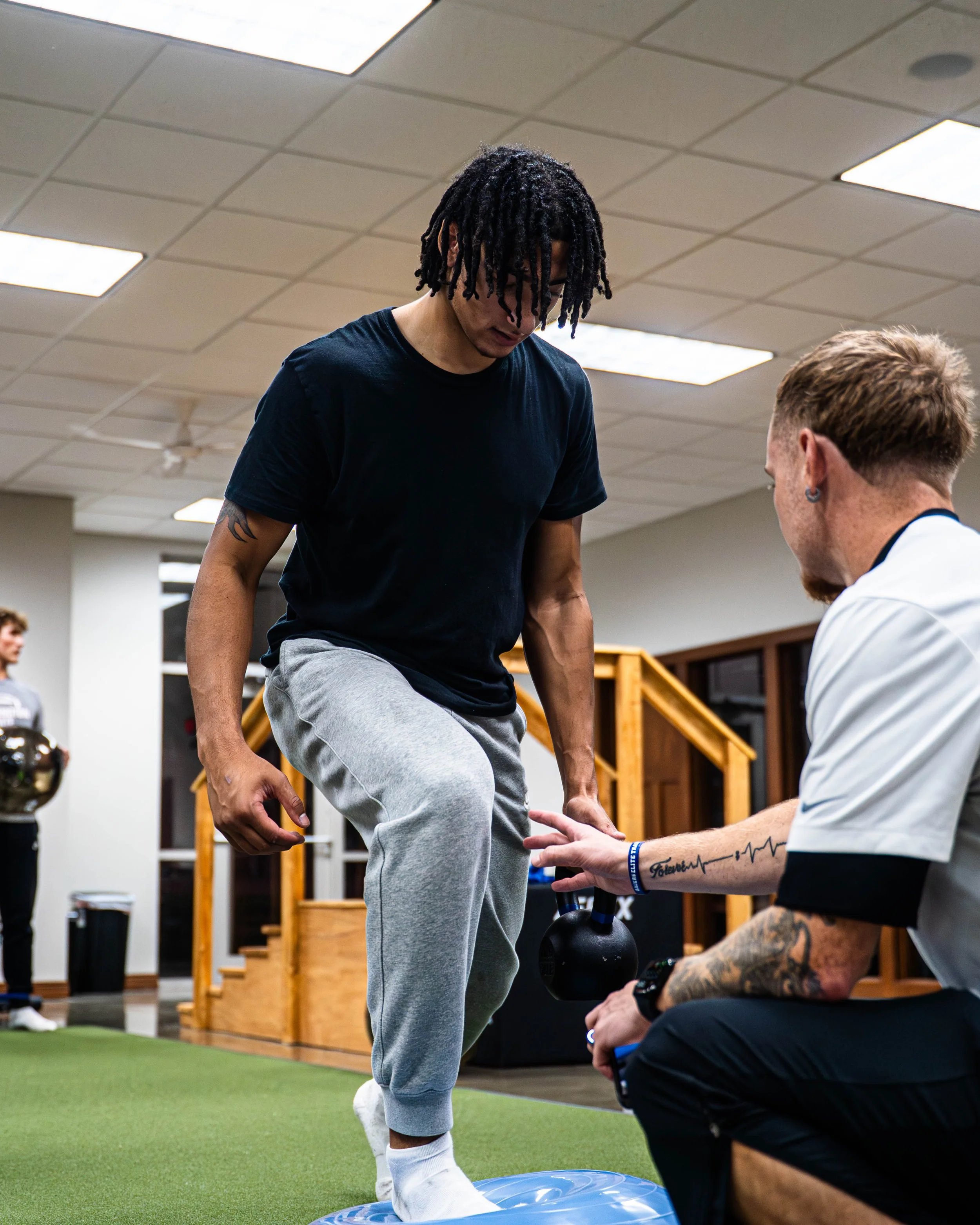 A man with dreadlocks in a black t-shirt and gray sweatpants balancing on a blue balance trainer during a physical therapy session with a therapist assisting him in a gym.