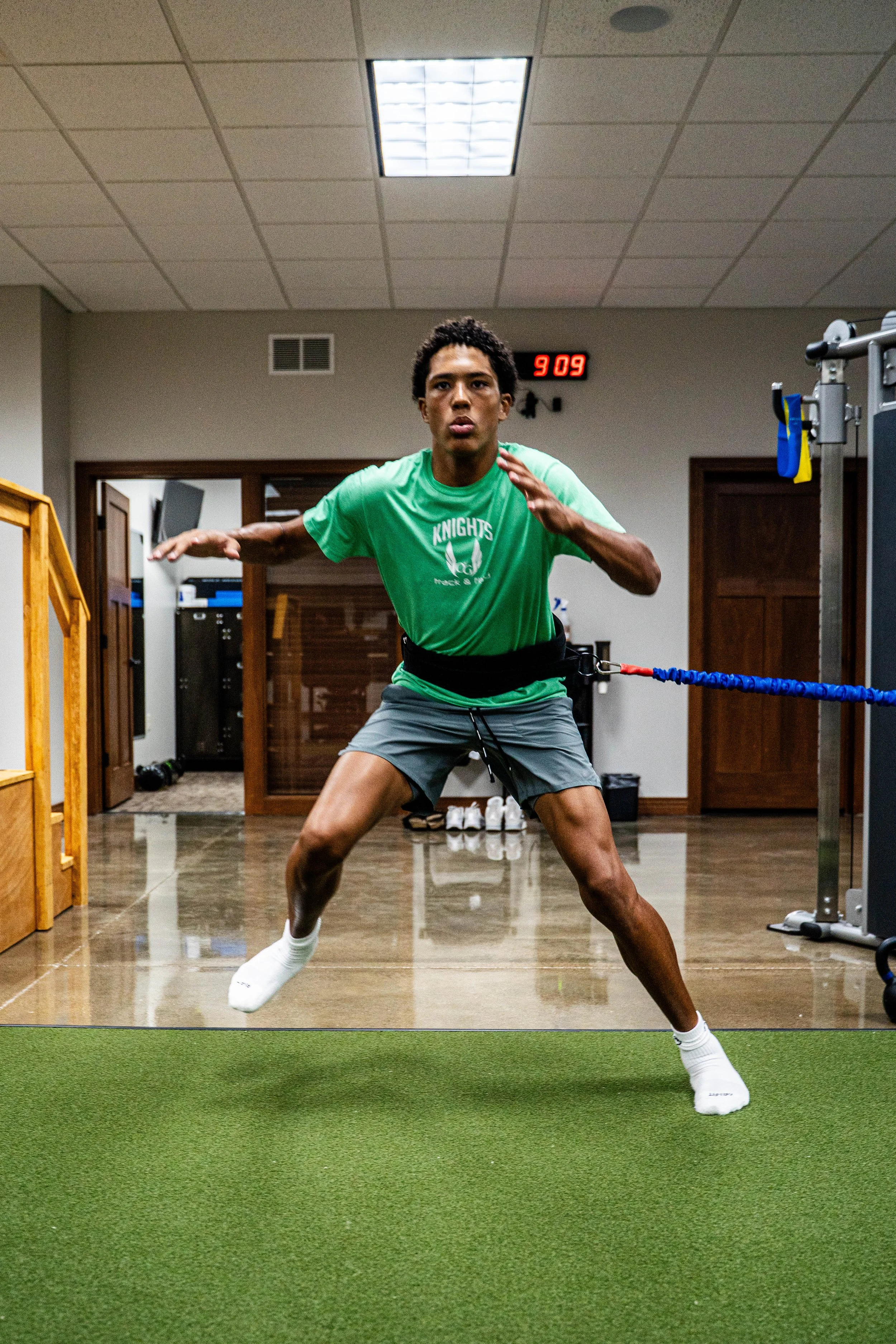 Young athlete performing resistance training with a blue resistance band attached to a wall-mounted apparatus in a gym.