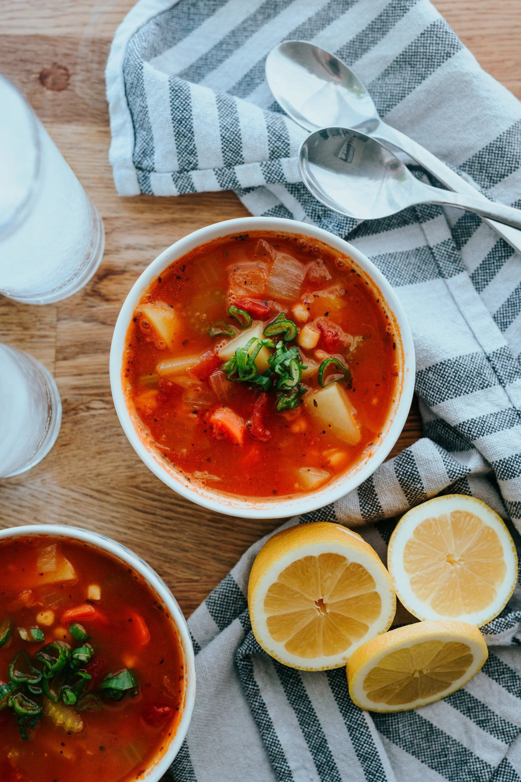 Bowl of tomato-based vegetable soup garnished with chopped green onions, with three lemon halves nearby, on a wooden table with a striped cloth, glass of water, and silver spoons.