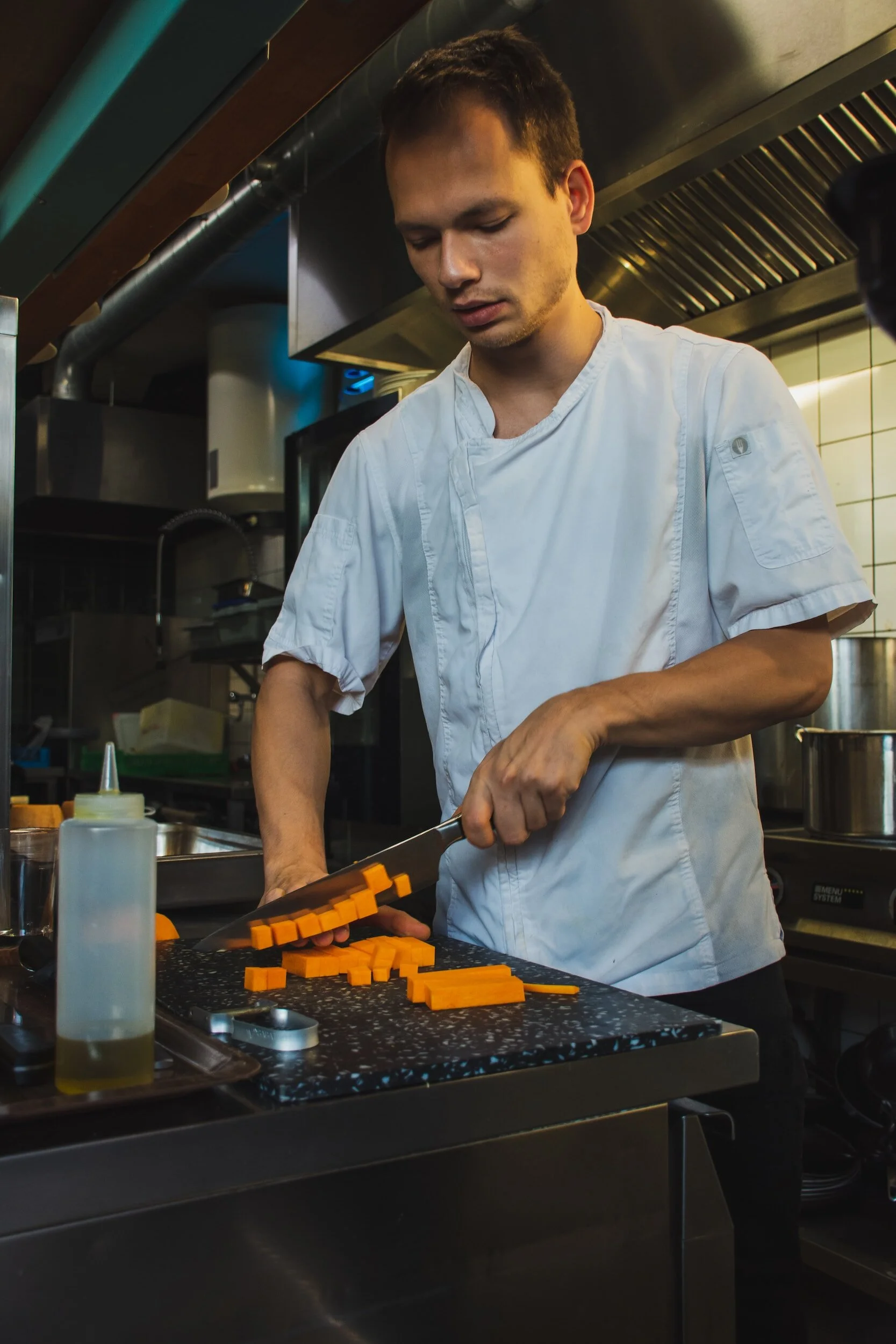 A chef chopping orange vegetables, likely carrots, on a black cutting board in a professional kitchen.