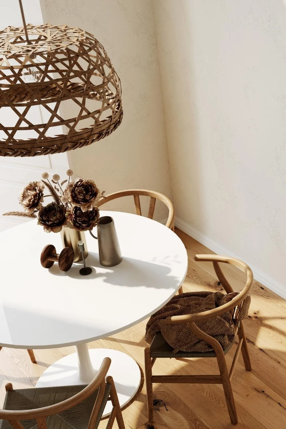 A round white table with a vase of brown artificial flowers and two wooden chairs in a bright room with light wood flooring and a woven pendant lamp overhead.