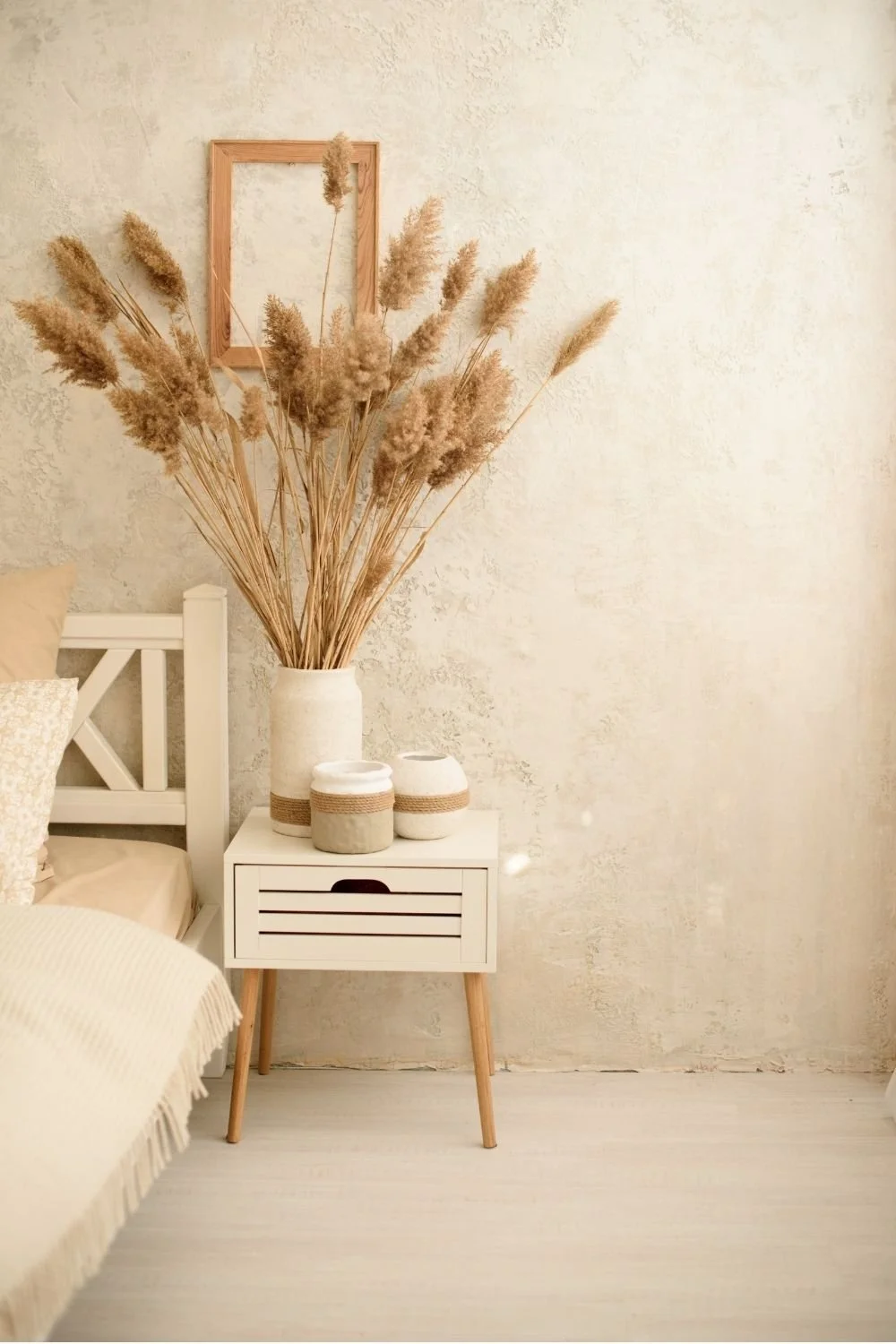 A bedroom corner with a white bedside table holding a large vase and three smaller containers, a bed with beige and white pillows, and dried pampas grass, against a textured white wall.