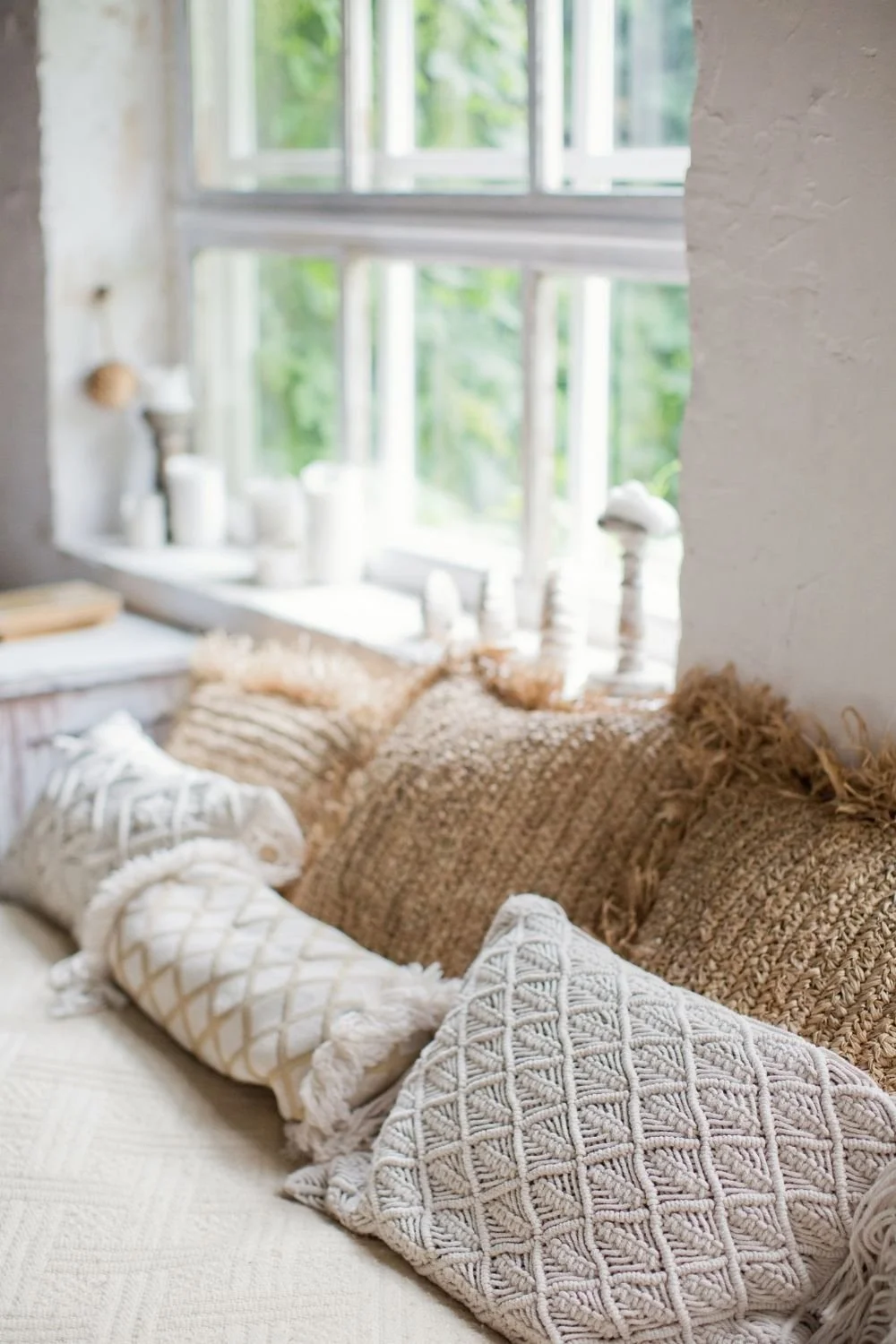 A cozy living room corner with beige and white decorative pillows on a cushioned window seat, overlooking a bright window with green foliage outside.