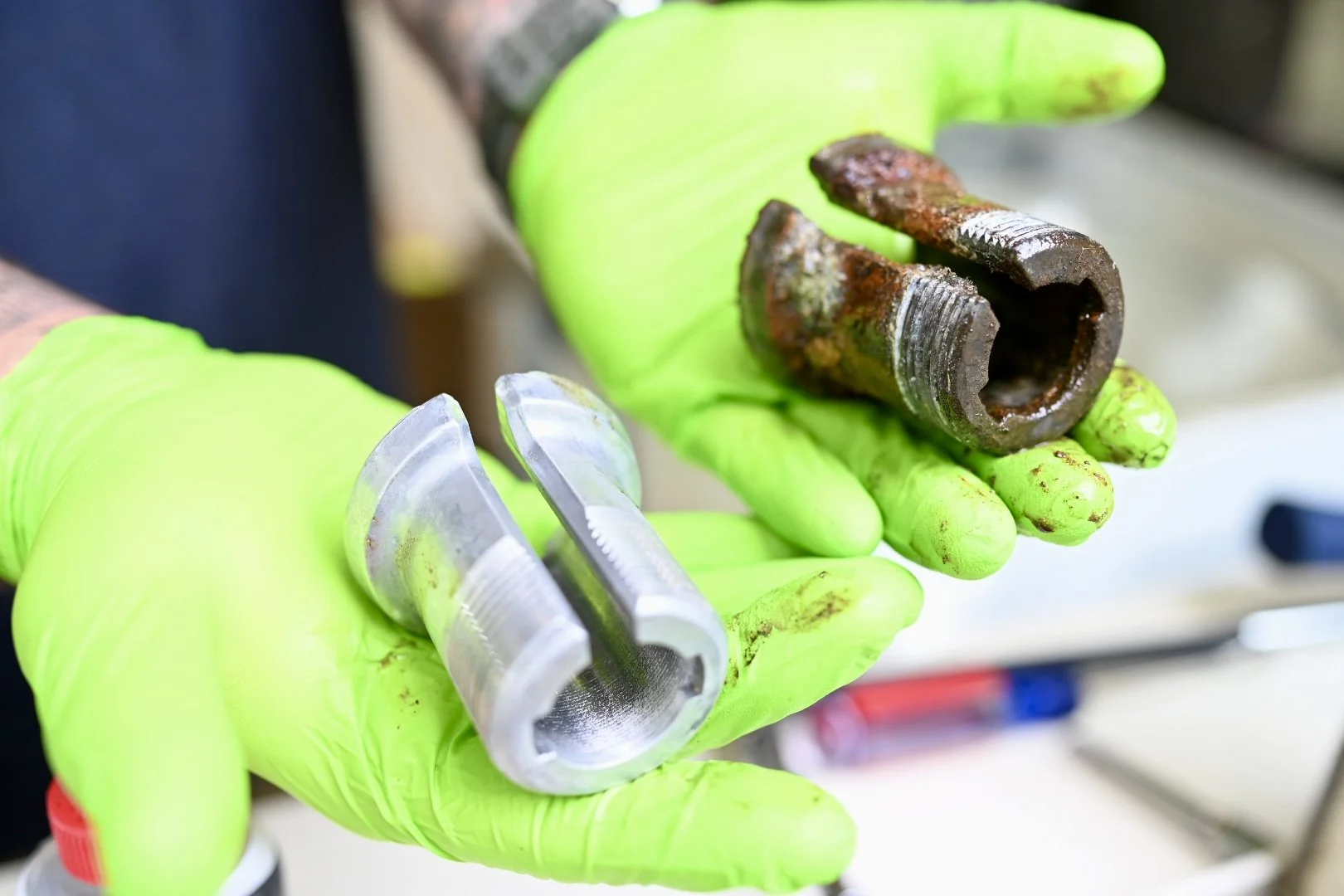 Person wearing bright green gloves holding a rusty, broken metal fitting and a shiny, new metal fitting.