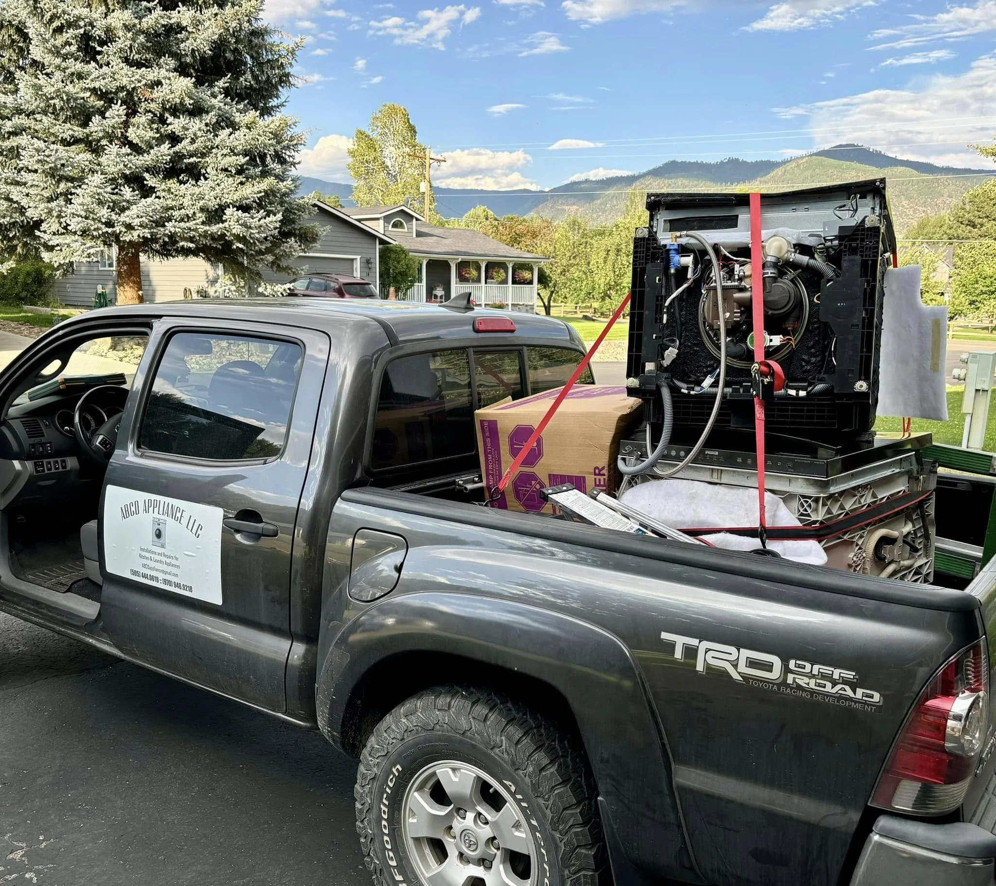 A gray Toyota Tacoma pickup truck with a sign on the door that reads 'ACD Appliance LLC' is parked outdoors in a residential area with houses, trees, and mountains in the background. The truck bed contains a large refrigerator secured with straps.