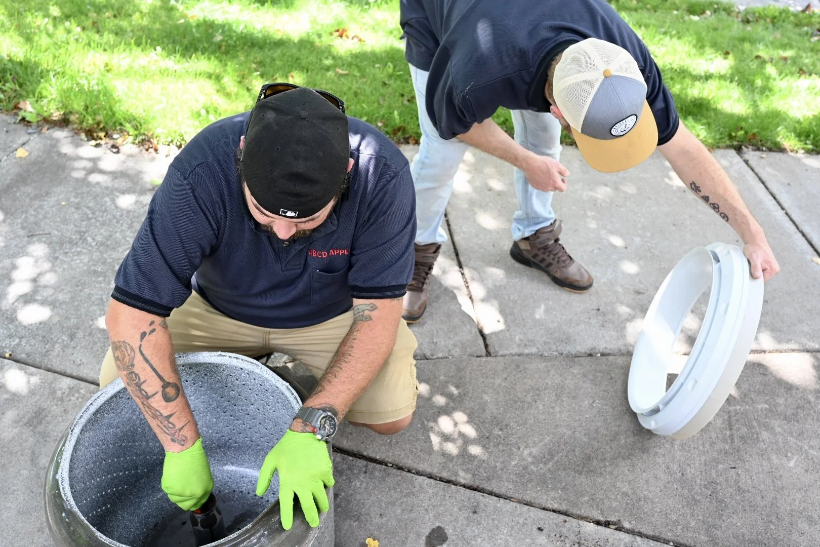 Two men working on cleaning washer parts on the sidewalk, with greenery and trees in the background.