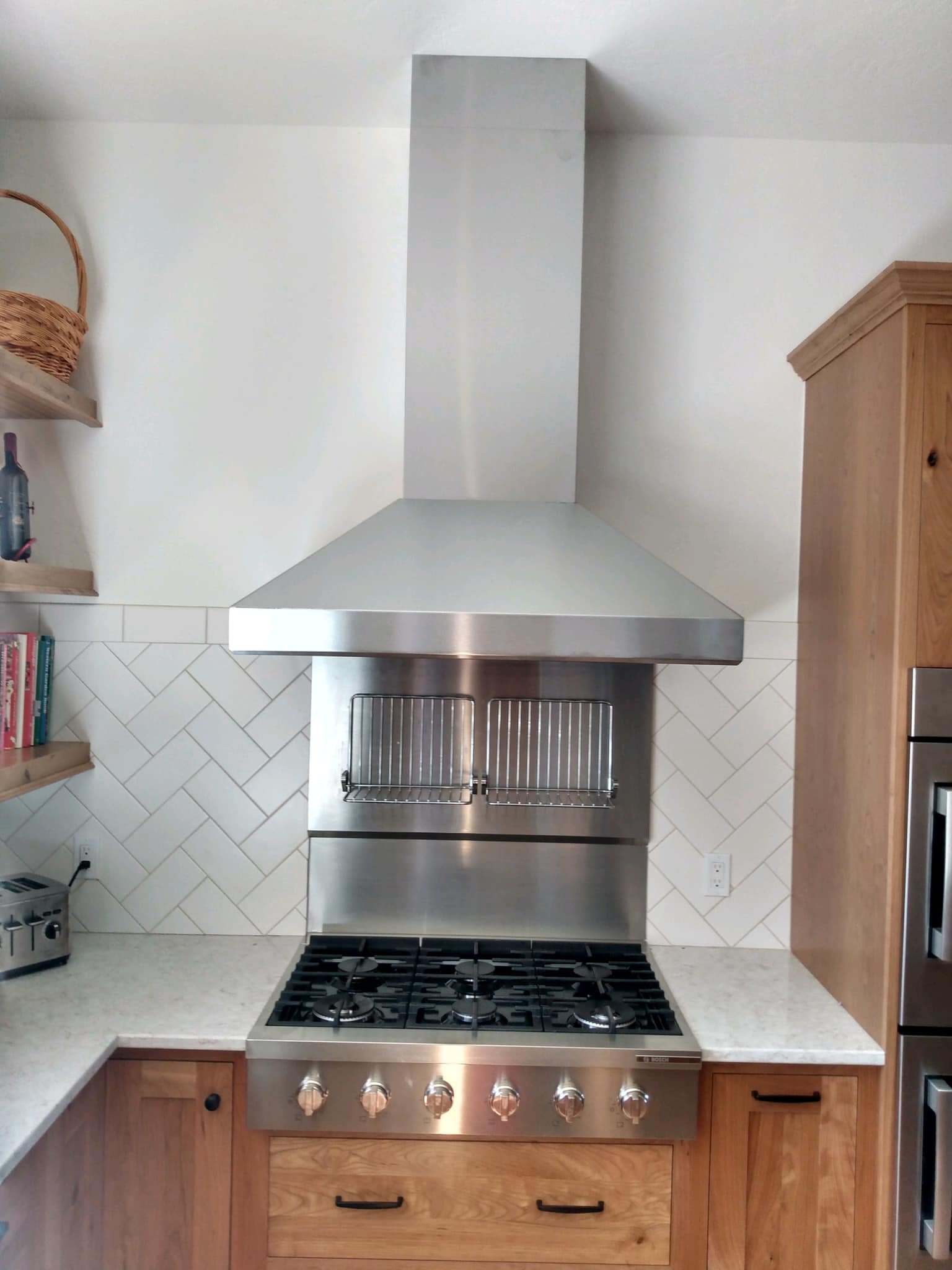 Modern stainless steel kitchen range with five burners, oven controls, and a versatile ventilation hood, flanked by wooden cabinets and open shelves with books and a basket.