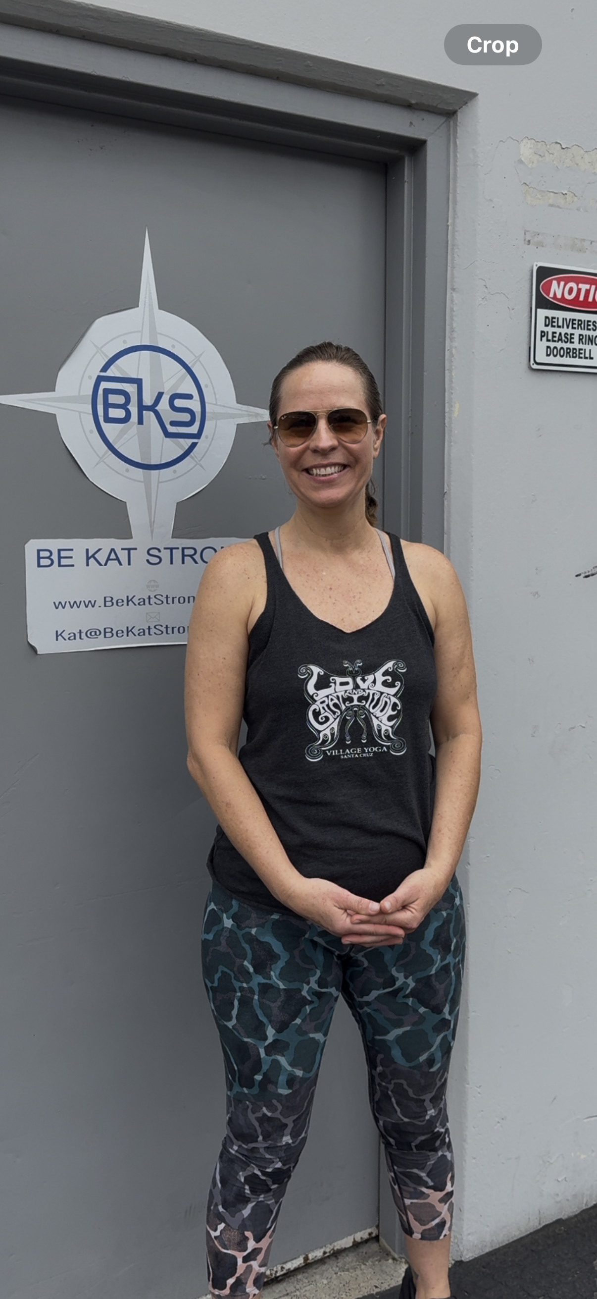A woman wearing sunglasses, a black tank top with a design that says "Love Created" from Village Yoga, and patterned leggings, standing in front of a gray door with a BKS logo and a sign that says "Be Kat Strong".