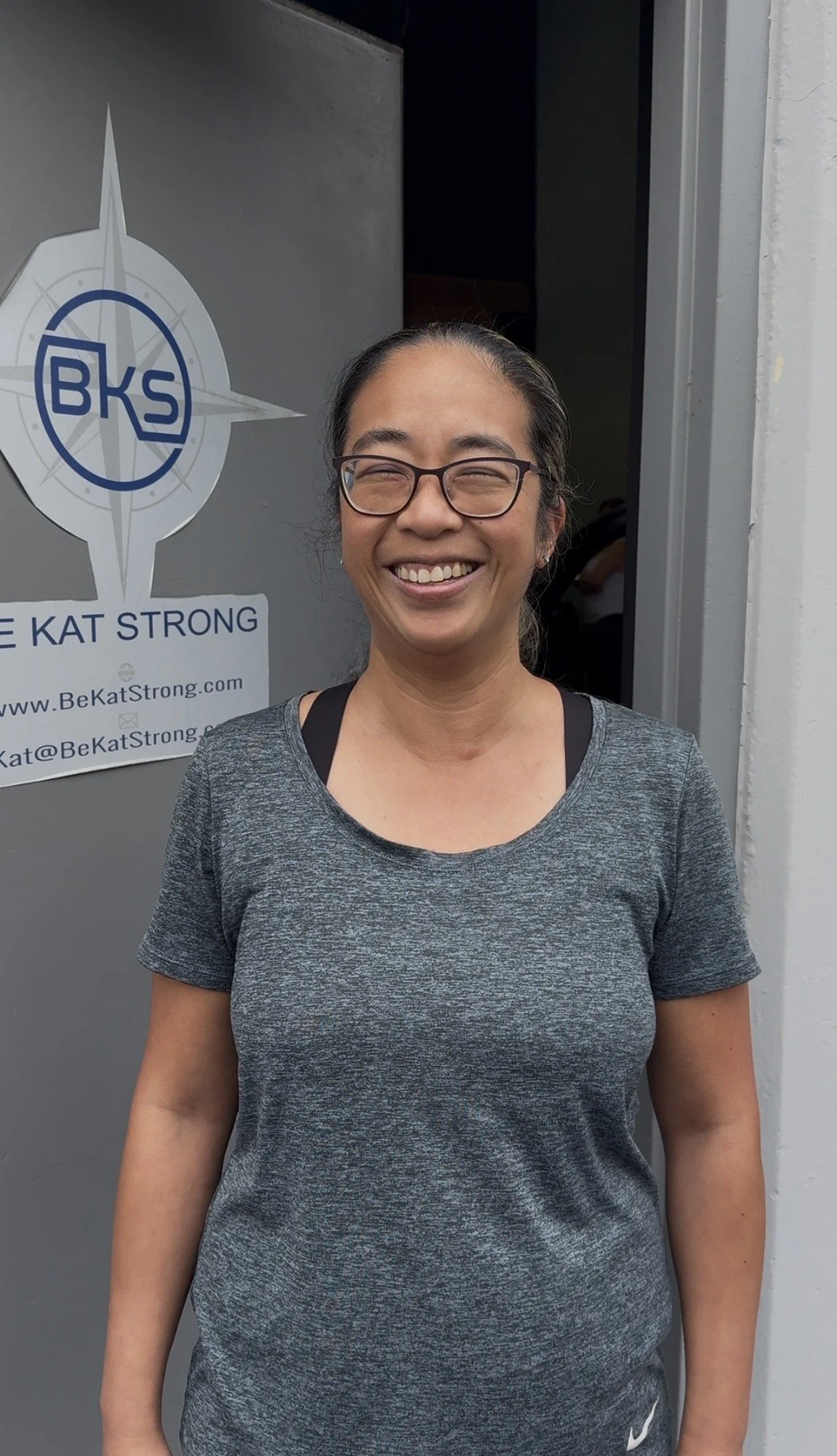 A smiling woman wearing glasses and a gray athletic shirt standing in front of a gray door with a logo and text for Be Kat Strong.