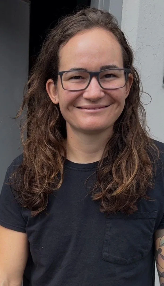 A woman with long, wavy brown hair and glasses, smiling, wearing a black T-shirt, standing outdoors near a gray wall.