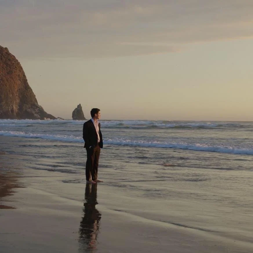 A photo of me in a black suit standing looking to the far distant ocean. A giant rock, cannon beach can be see behind. Low soft waves crashing near by.