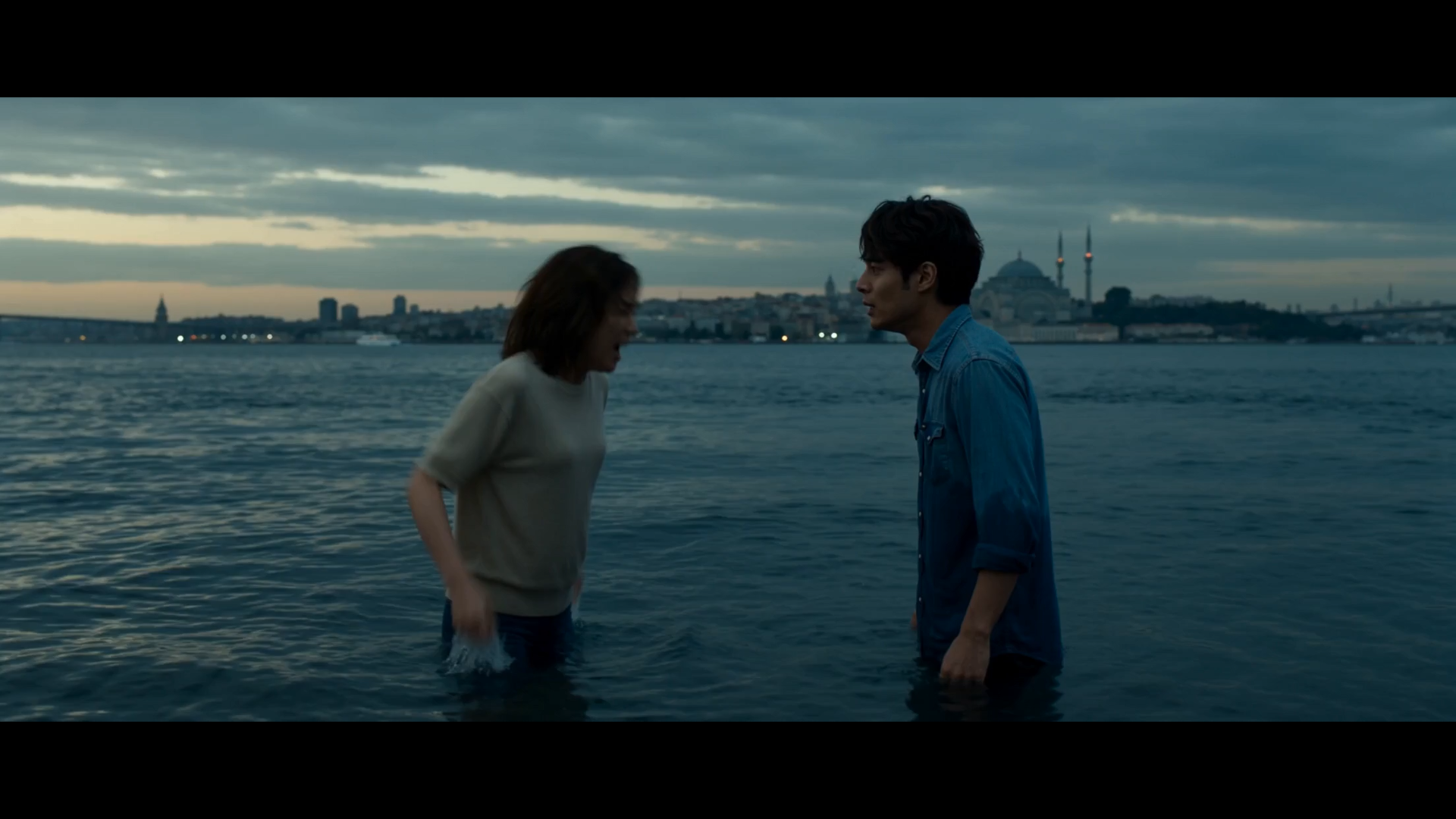 Two characters standing in water beneath a cloudy evening sky, with a city skyline behind them.