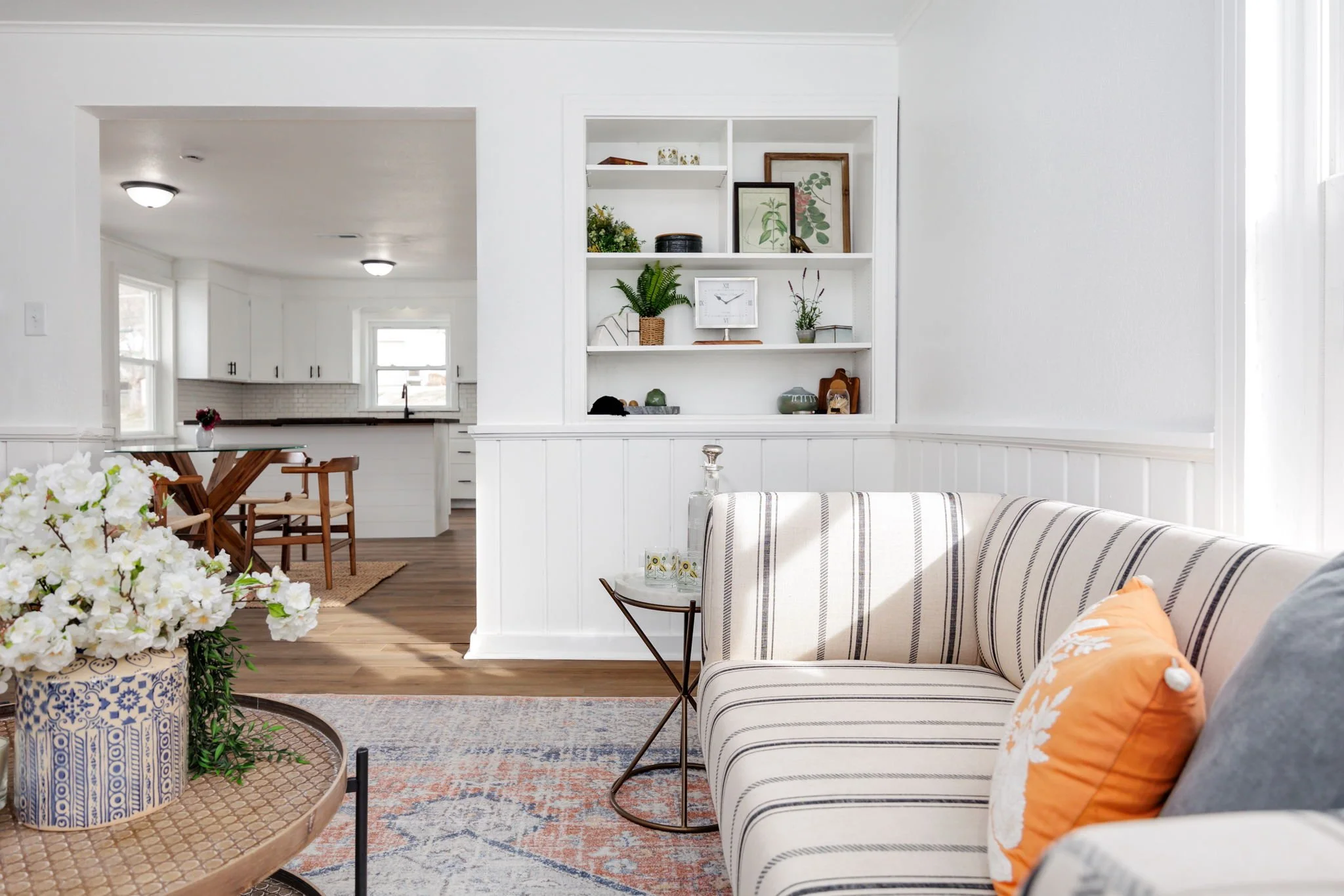 Bright living room with striped sofa, orange pillow, white flower arrangement in decorative vase, open shelves with decor, and view of a kitchen in the background.