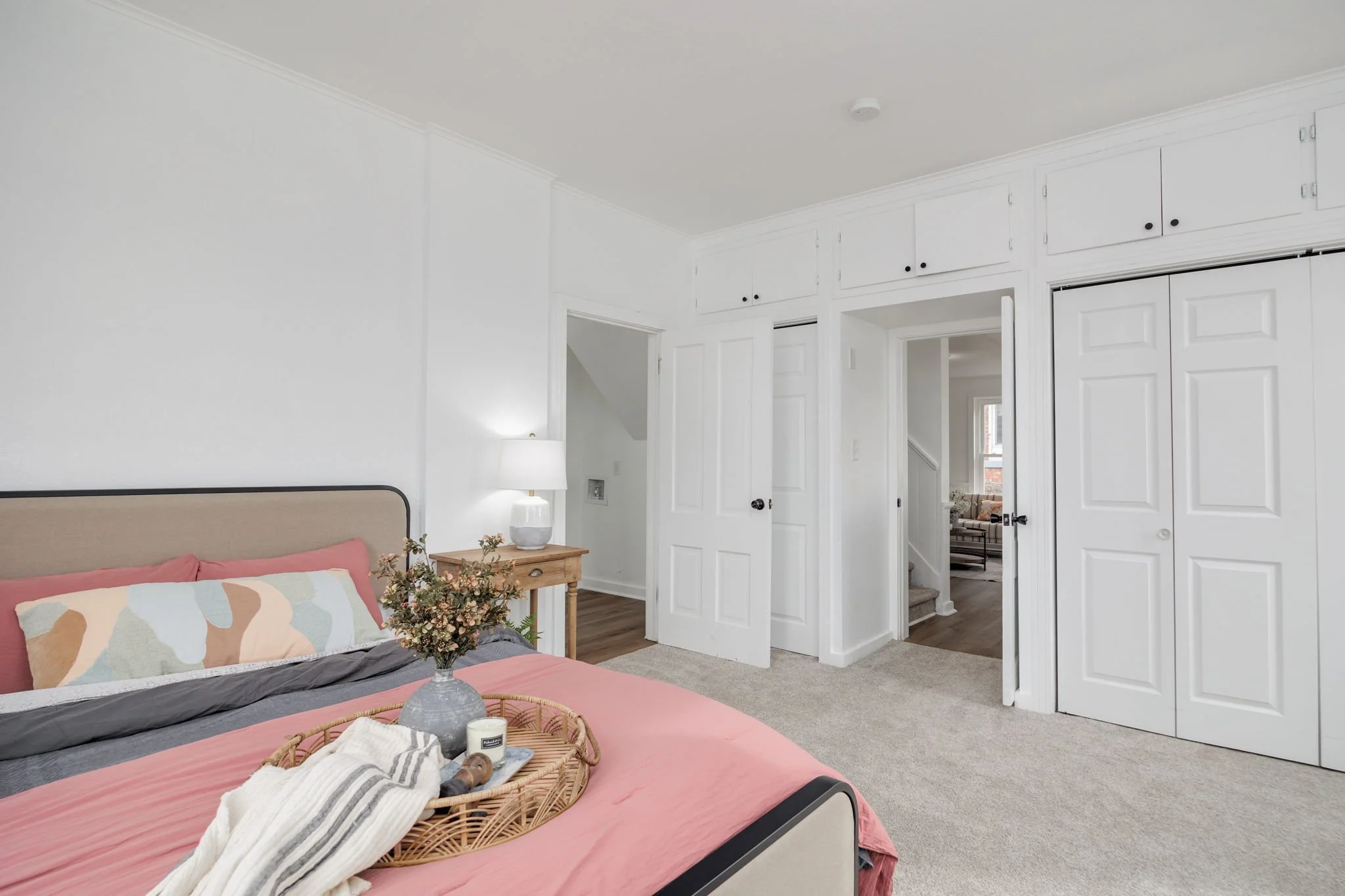 Modern bedroom with white walls, pink bedding, decorative pillows, and a wood side table with a lamp. Basket with items on bed. White doors leading to hallway.
