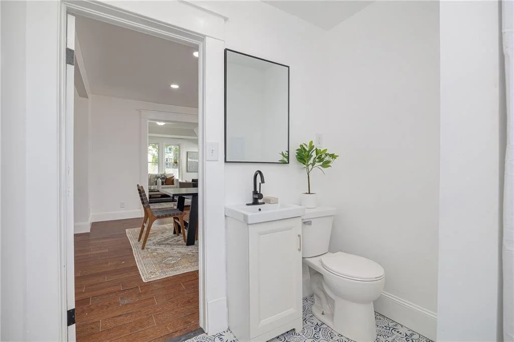 Modern bathroom with a white vanity, black faucet, rectangular mirror, potted plant, toilet, and patterned floor tiles. Open door leads to a living room with wooden flooring and a dining table.