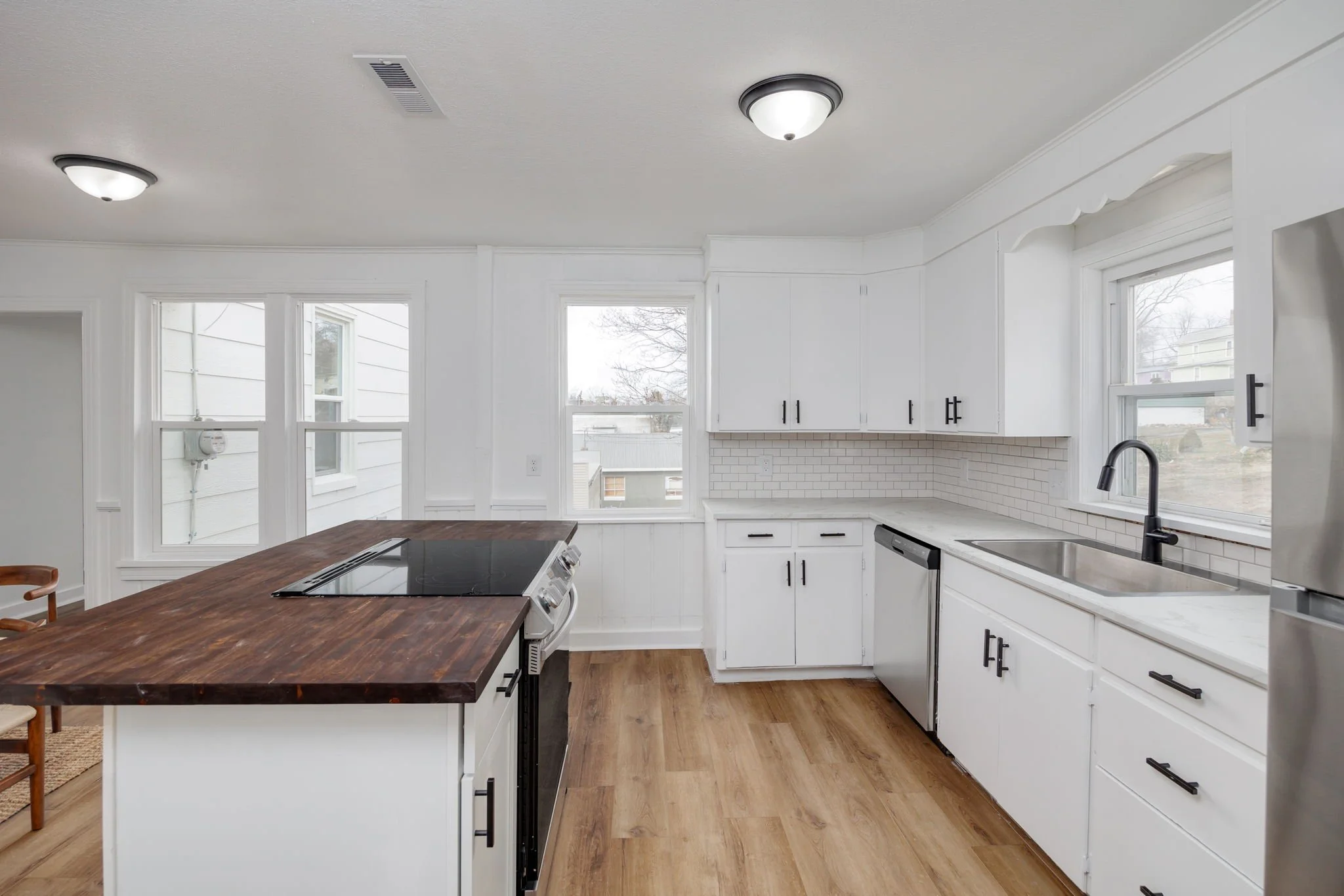Modern kitchen with white cabinets, wood countertop, stainless steel appliances, black faucet, and hardwood flooring. Large windows provide natural light.