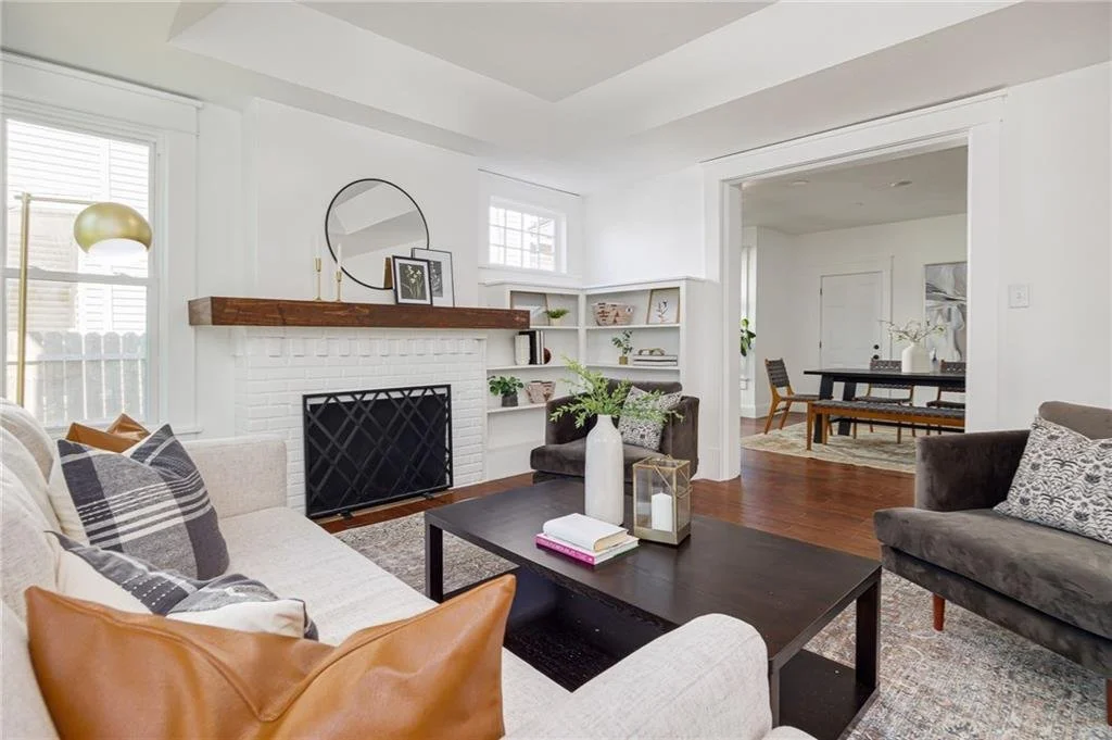 Modern living room with white walls, a couch with pillows, a black coffee table, and a white brick fireplace adorned with a round mirror and decor. Wooden mantle, bookshelves, and a glimpse of a dining area with a wooden table and chairs in the backg