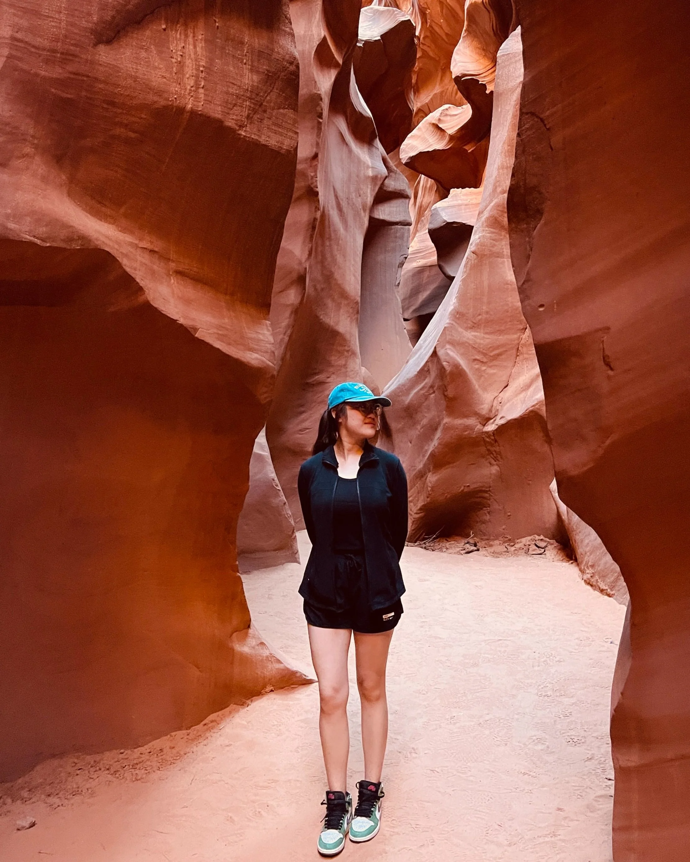 A woman standing in a narrow sandstone canyon, wearing a black outfit, blue cap, and sneakers.