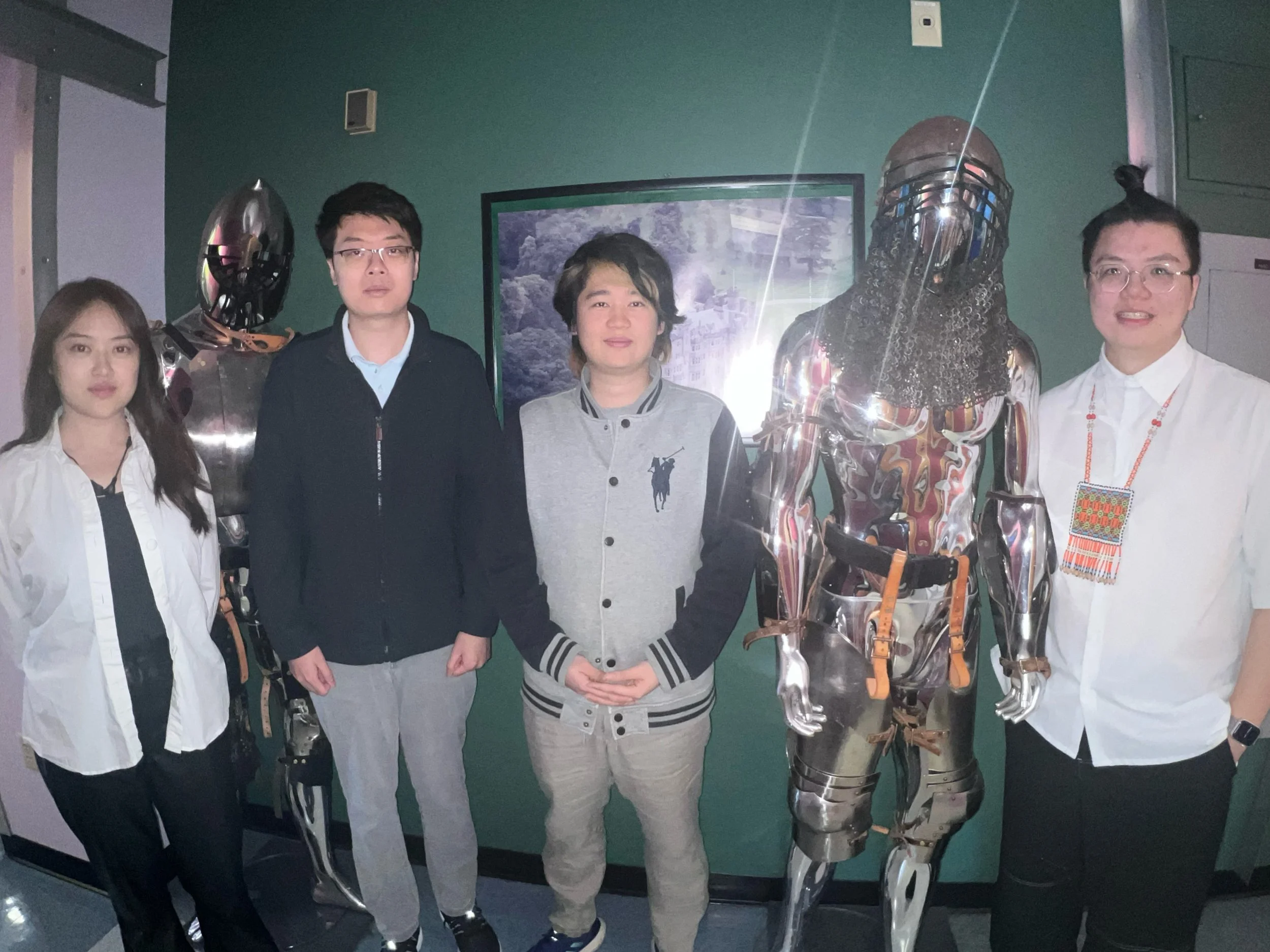 Five people standing with two humanoid robots, one with a reflective metallic surface and the other with a complex robotic suit, in a room with a green wall and a framed picture.