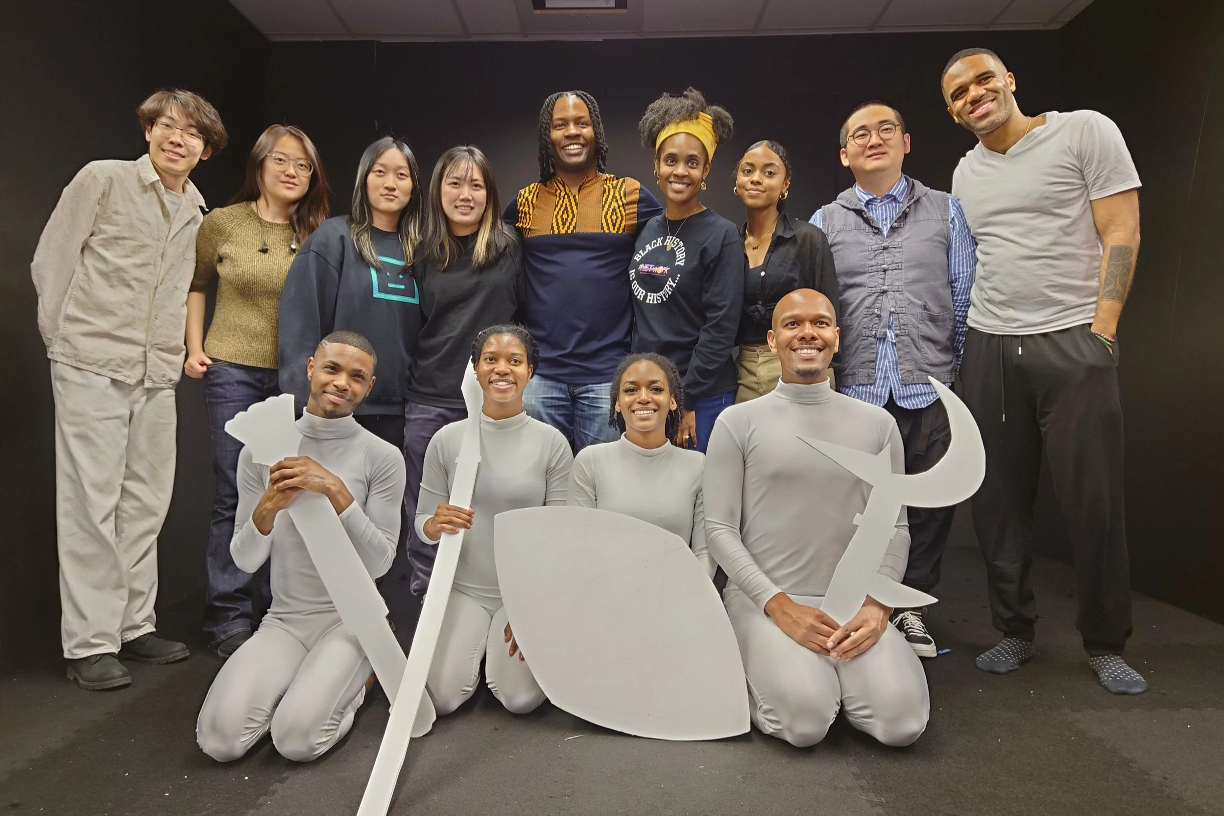 Group of diverse people, including men and women, posing together in a black-walled room, some kneeling in front holding white cardboard cutouts of a tree, a leaf, and a spear.