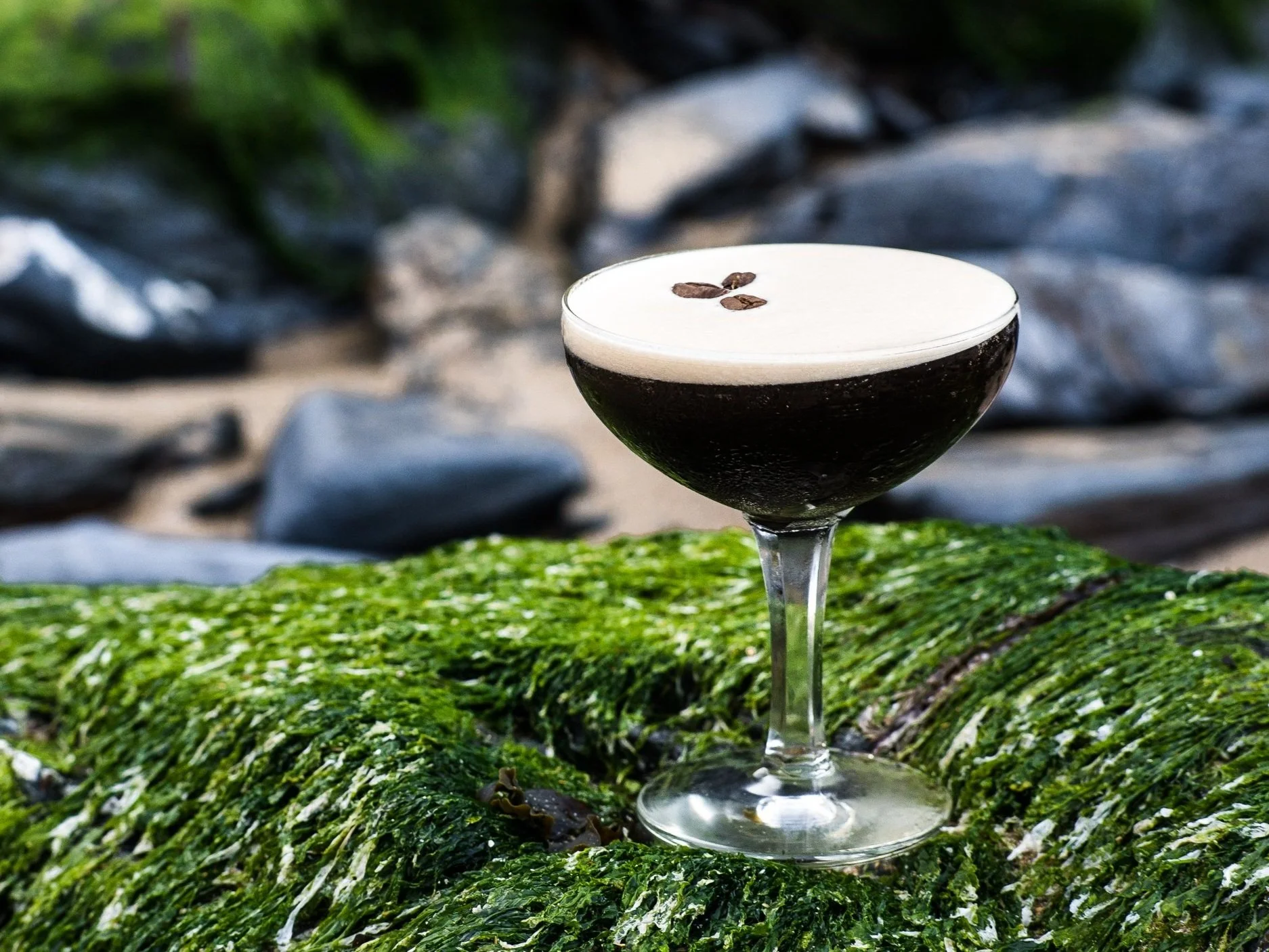 An atlantic distillery espresso martini cocktail with a frothy white top garnished with coffee beans, placed on a mossy rock near a rocky beach.