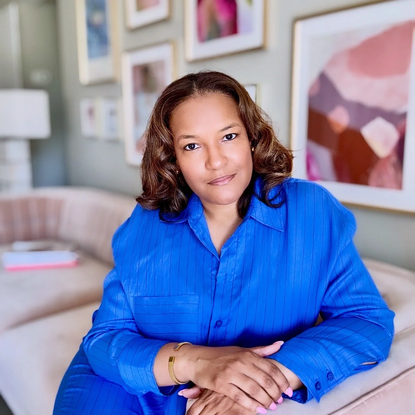Aisha Holly seated in a bright studio space surrounded by colorful artwork and framed prints, wearing a cobalt blue jumpsuit, hands resting in her lap, direct and warm gaze.