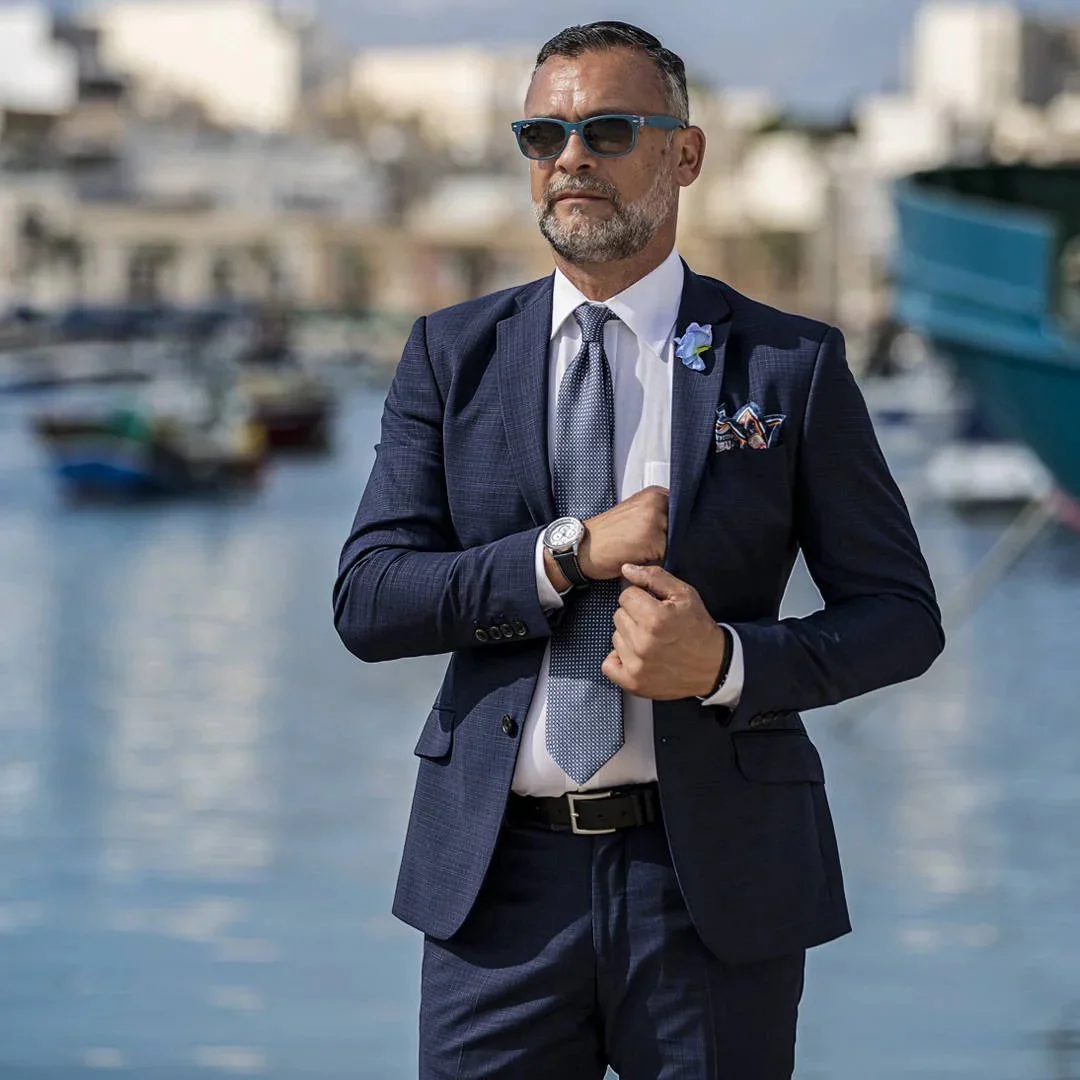 A man wearing a navy suit, white shirt, and gray tie, standing near boats in a marina, with sunglasses and a watch.