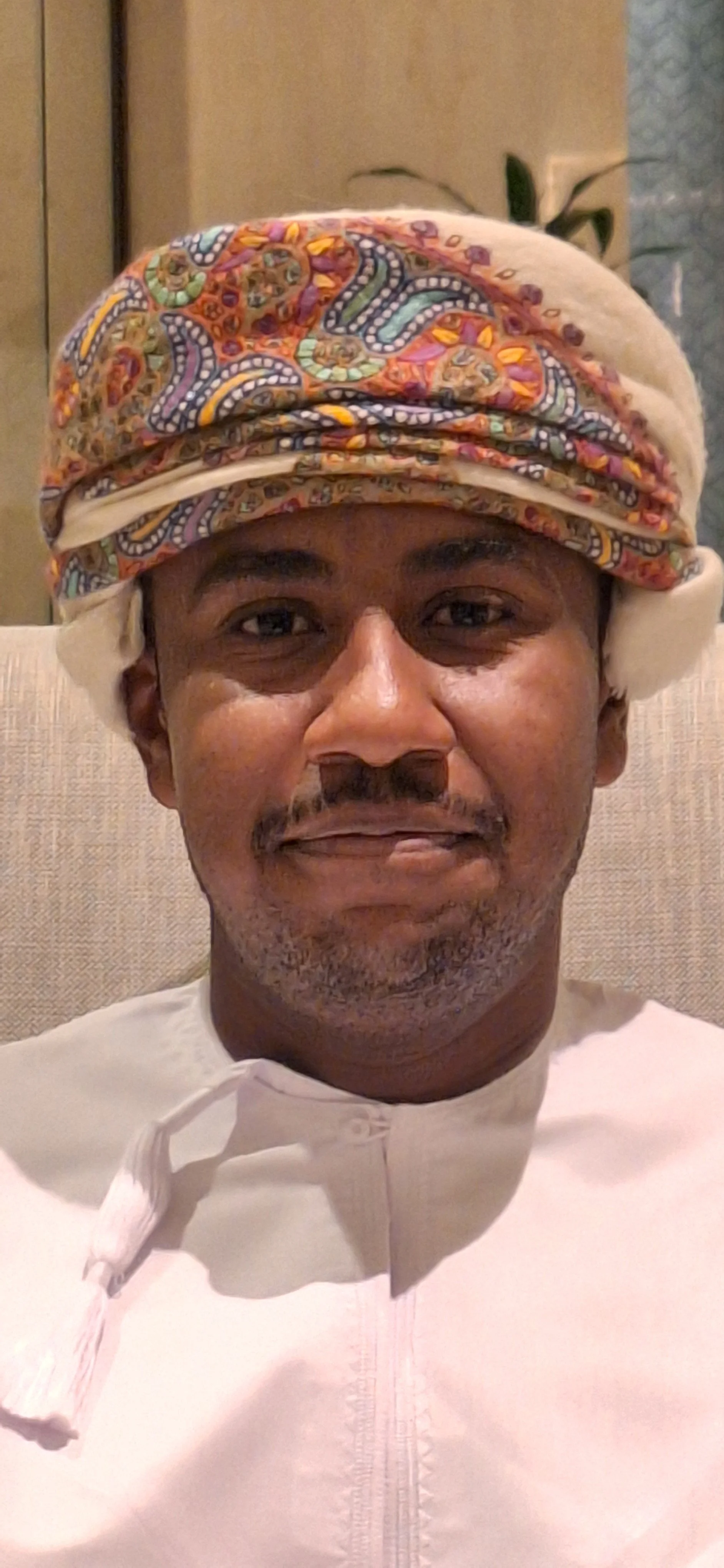 A man wearing a white traditional garment and a colorful patterned headscarf, sitting on a beige chair indoors.