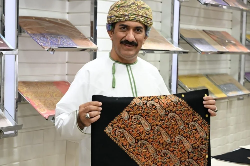 A man wearing traditional Omani clothing and a headscarf, standing in front of a display of fabrics, holding a piece of black fabric with intricate orange and gold embroidery.