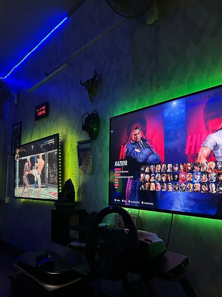 A gaming room with two LED screens on a wall displaying a fighting match and a character selection screen, surrounded by colorful LED lights, with various gaming memorabilia and equipment visible.