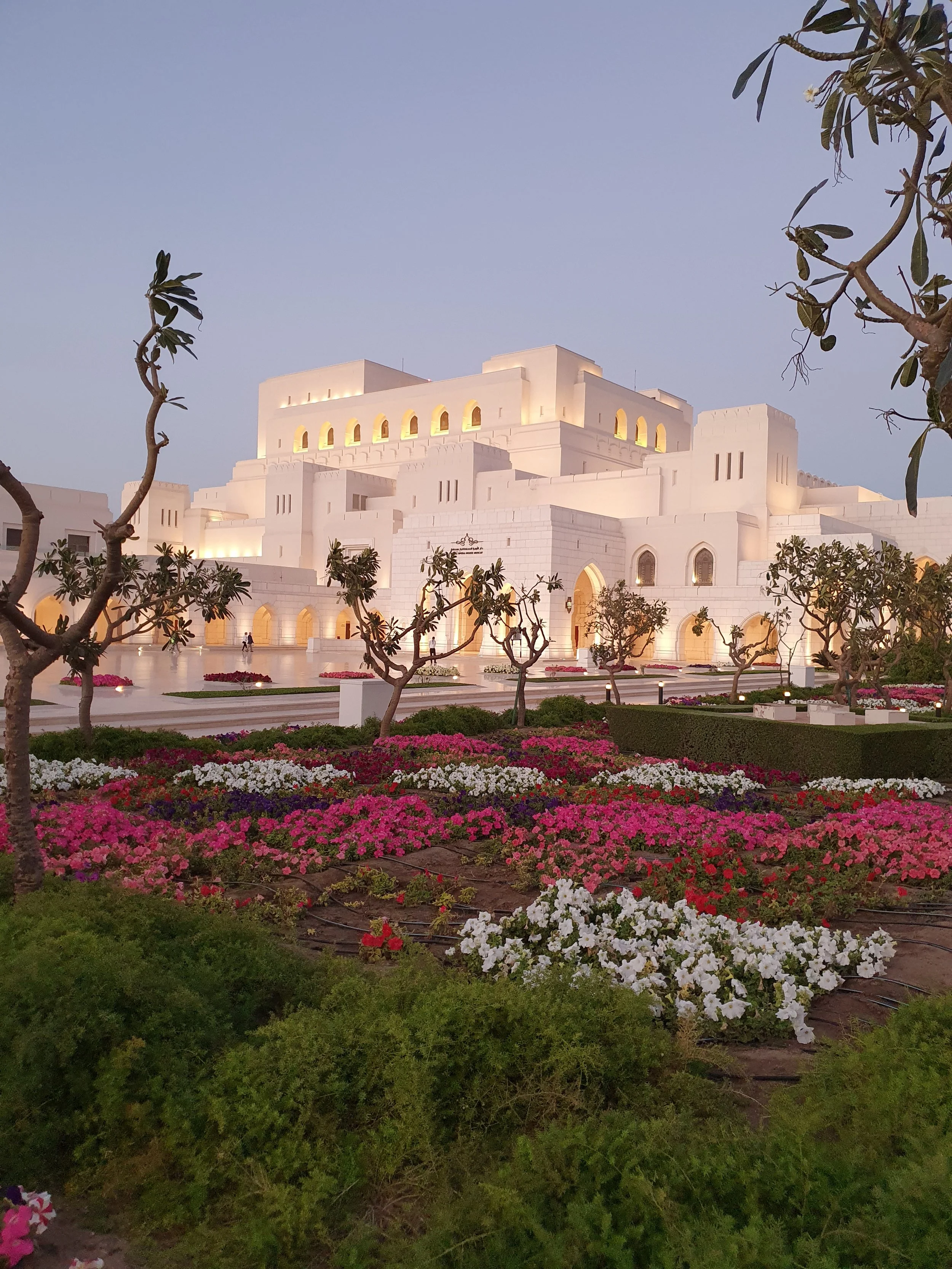 A large white building with a castle-like appearance, surrounded by colorful flower beds and trees, under a clear evening sky.