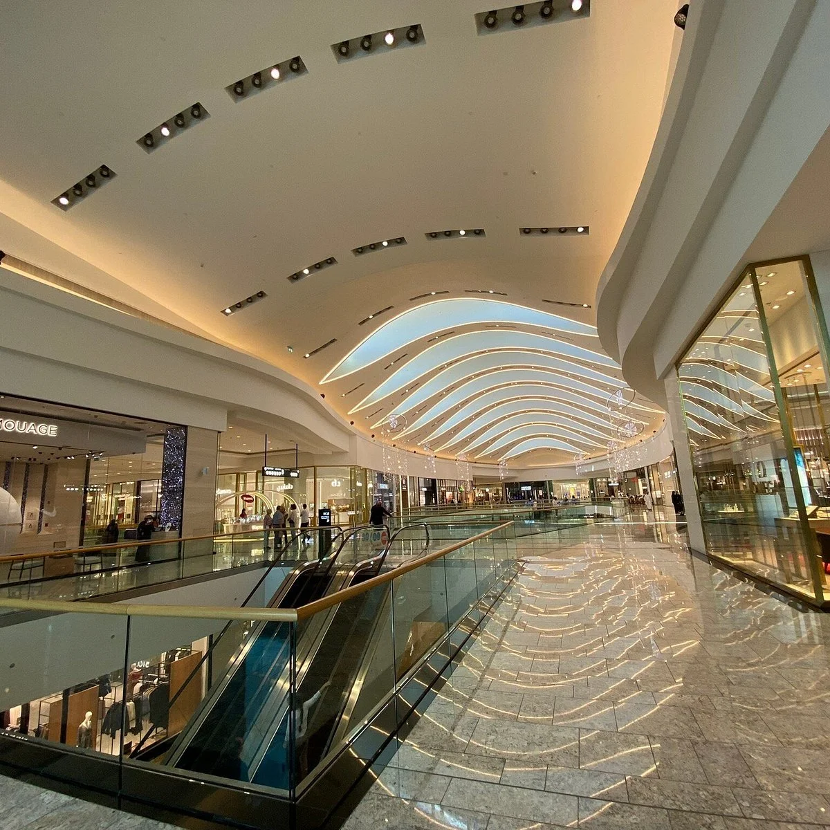 Interior view of a modern shopping mall with a curved ceiling design, escalators, and store fronts with glass windows.