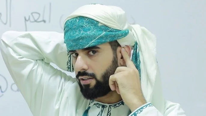 Man wearing traditional Middle Eastern attire, adjusting his headscarf, standing in front of a whiteboard.