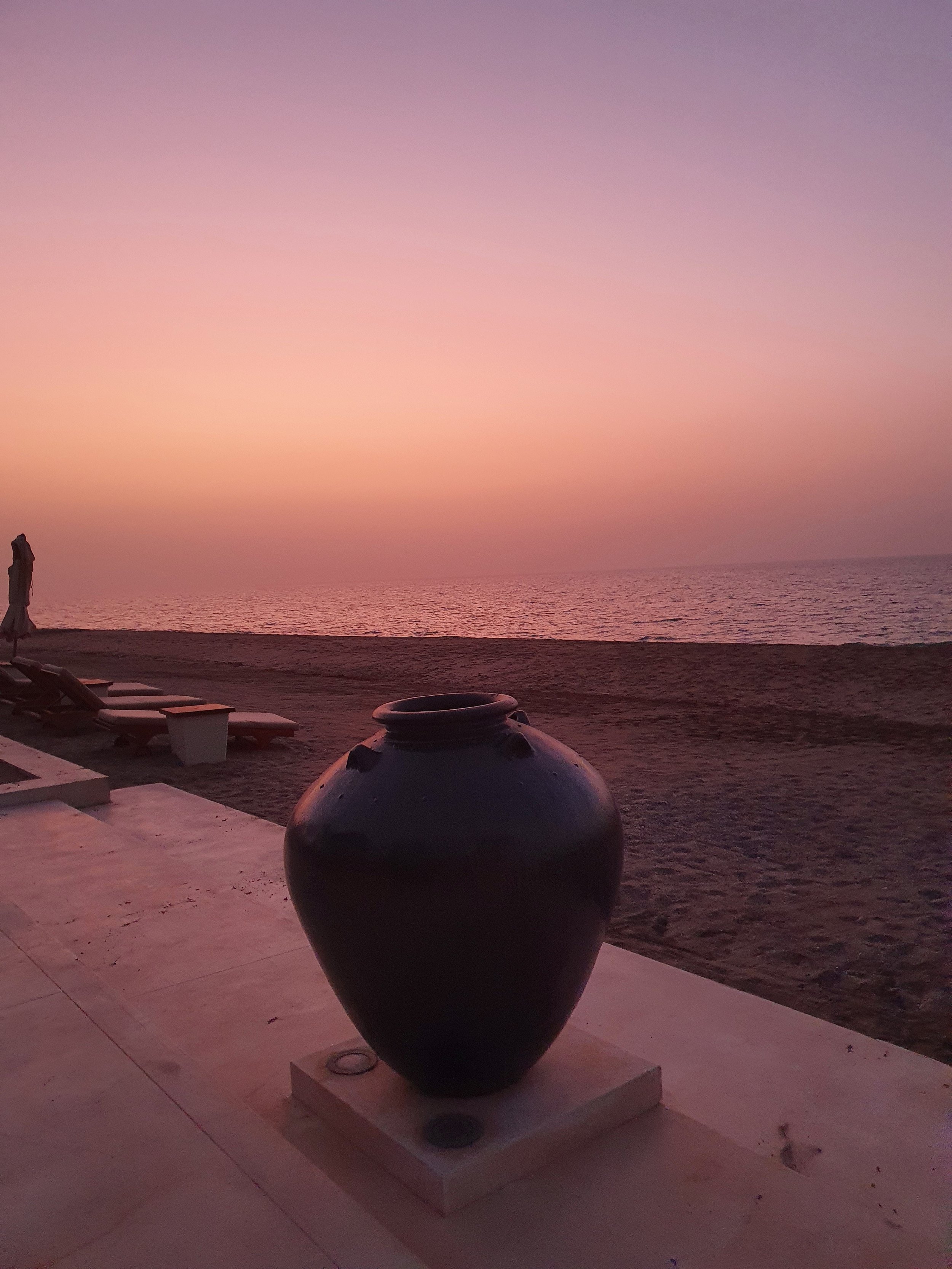 A large black vase on a white pedestal on a beachside patio during sunset, with lounge chairs and an umbrella in the background, overlooking calm ocean water and a pink-purple sky.