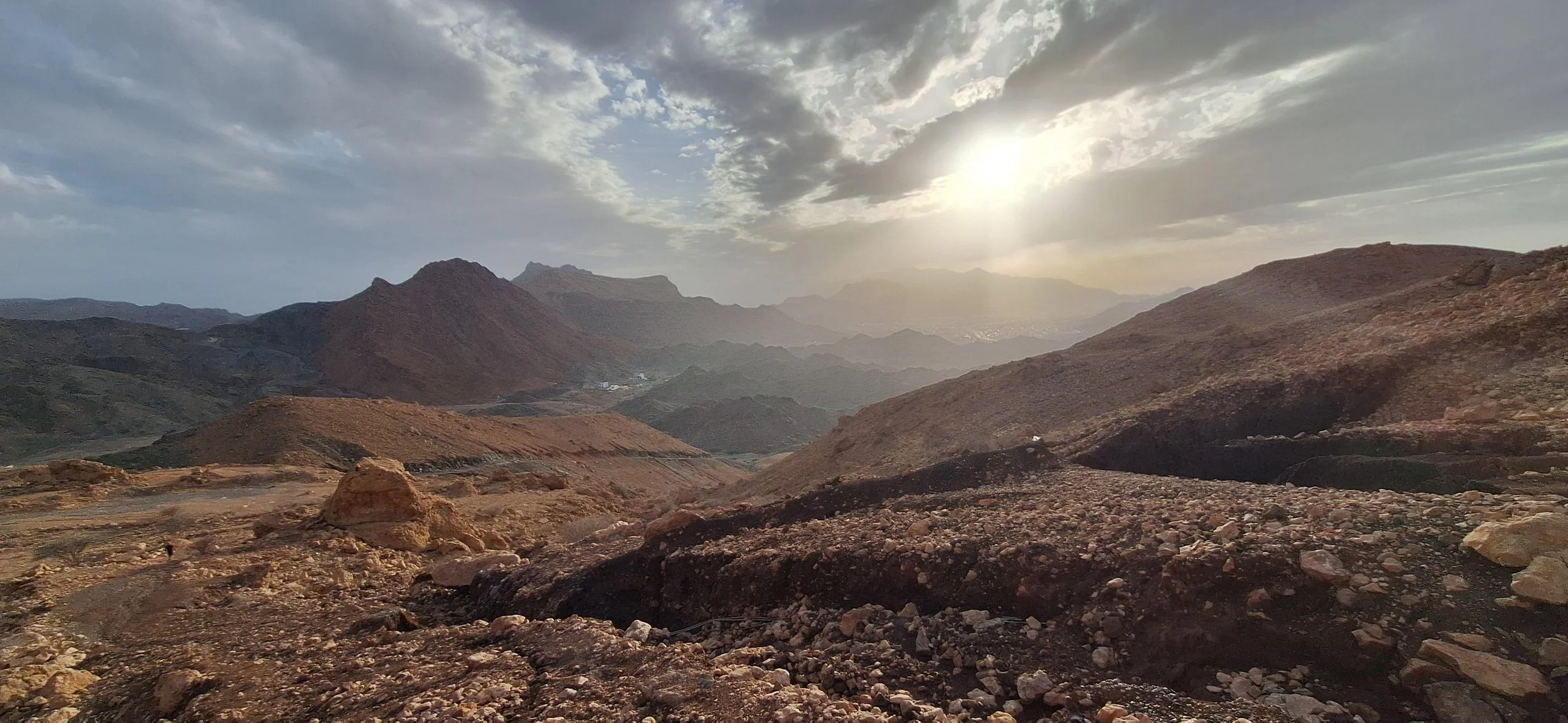 Sunset over rugged mountains with a rocky desert landscape in the foreground and partly cloudy sky.