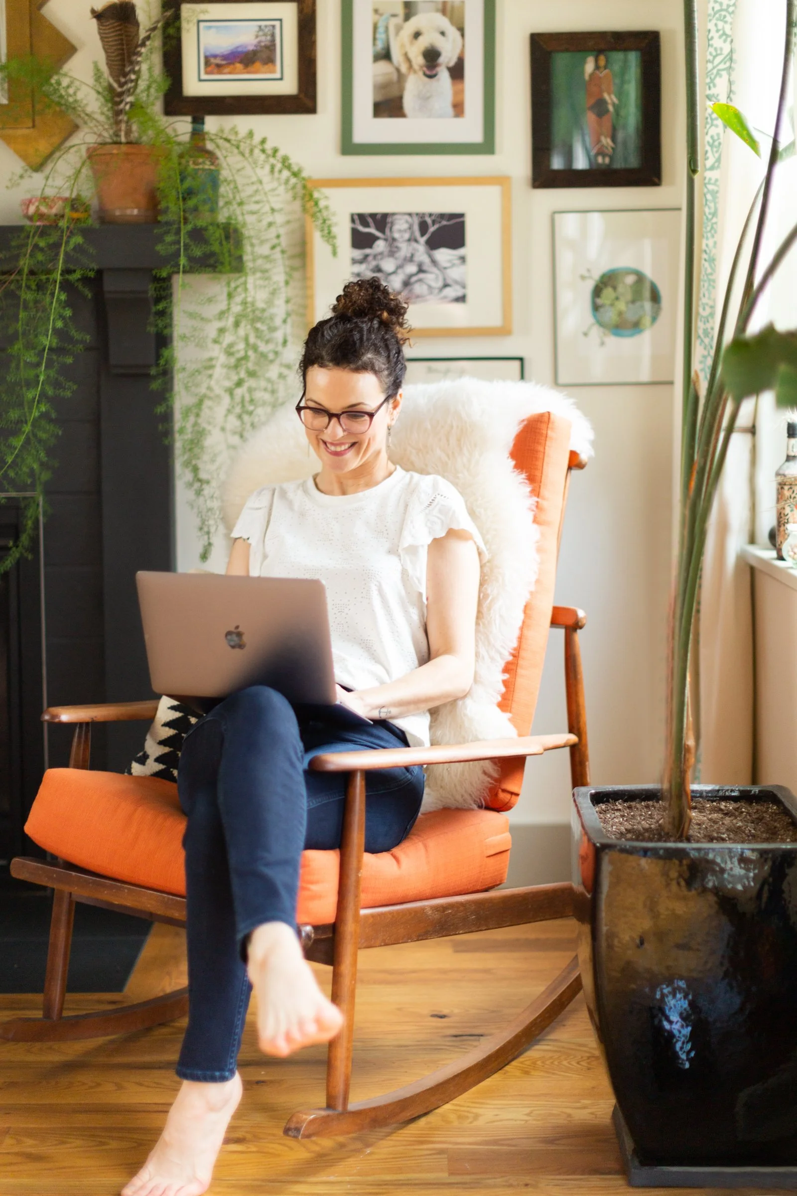 A woman with glasses and curly hair tied up in a bun sitting in an orange rocking chair, smiling while working on her laptop. The room has artwork on the wall, a large potted plant, and a wooden floor.