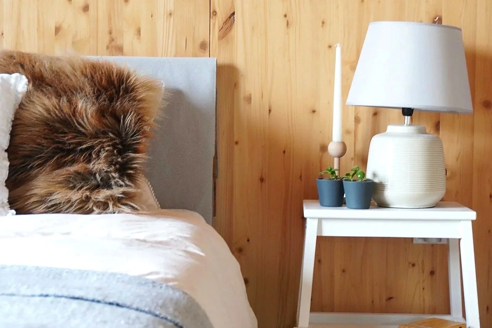Sunlit bed and table lamp in a small cozy home — natural wood cladded wall behind, simple bedside bench and small houseplants.