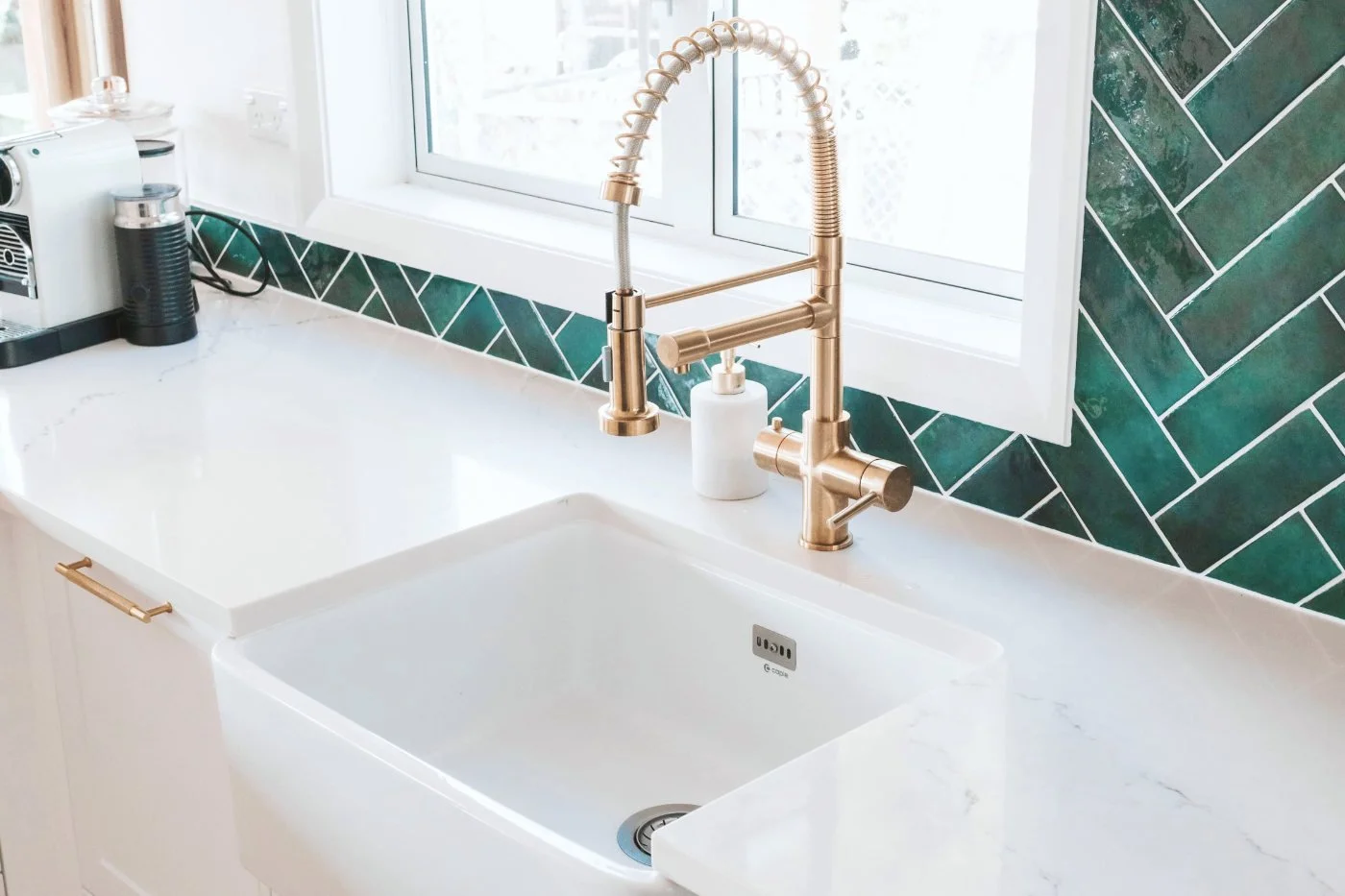 White kitchen sink under window with green tile backsplash and natural daylight.