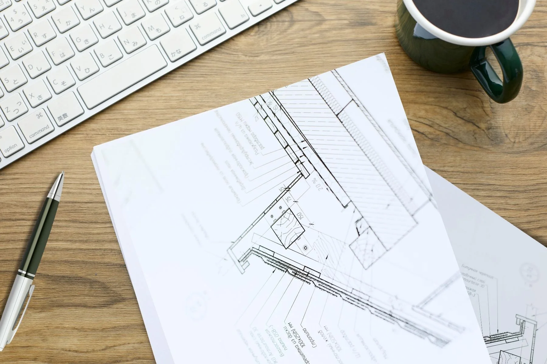 Top-down view of architectural plans, a pen, a keyboard, and a coffee mug on a table, illustrating eco-home design planning.