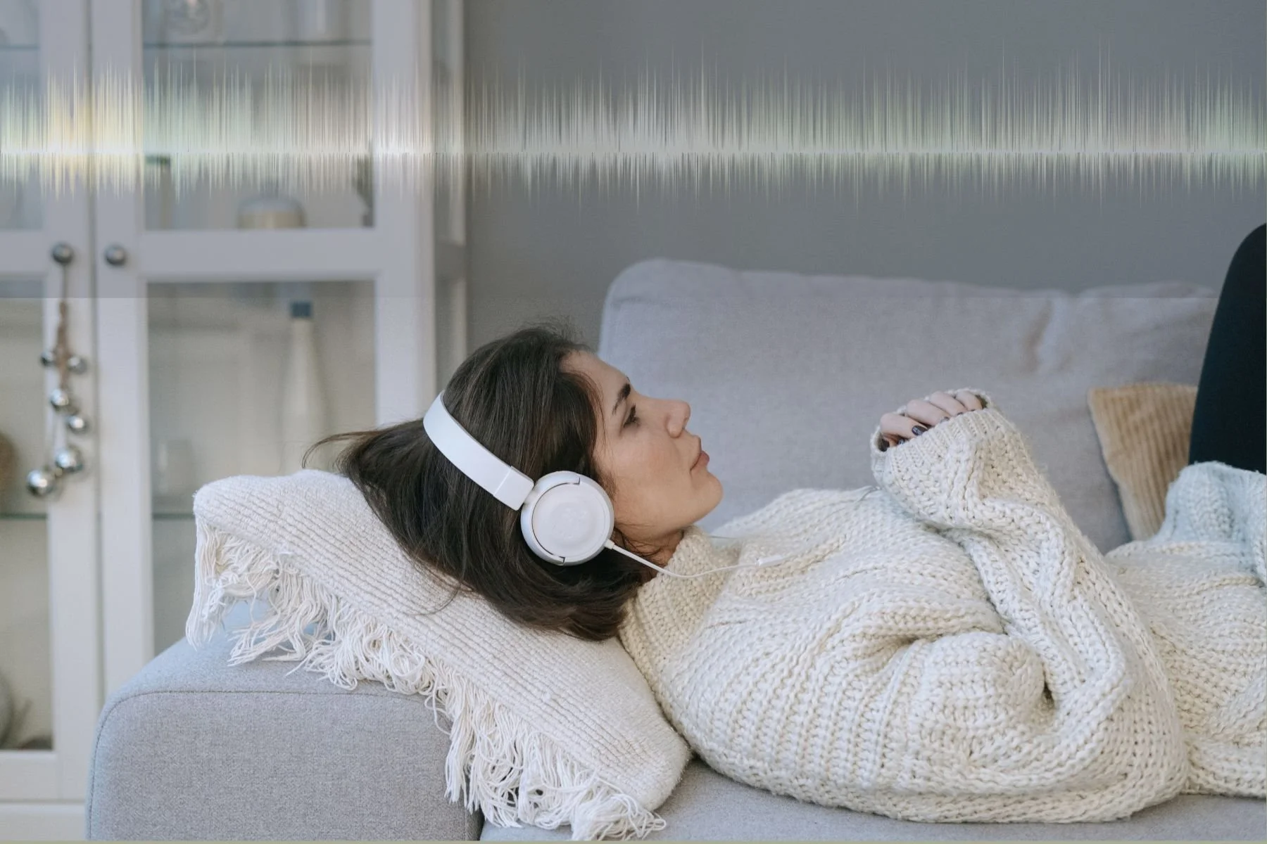 Woman lying down on the sofa listening to headphones and above her head a collage of sound waves representing sound comfort at home.