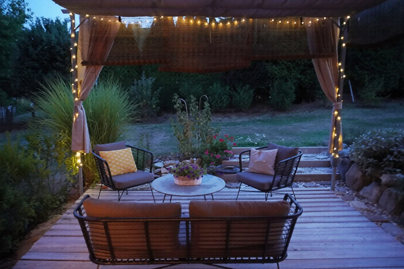 Cosy backyard patio design at night with warm twinkle lights, comfortable outdoor seating, potted summer flowers.