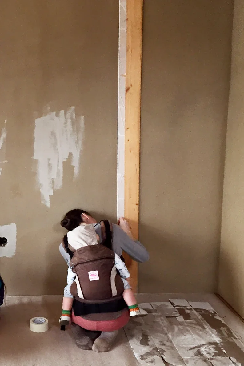 Woman preparing a clay plaster wall for painting while carrying a baby during home construction