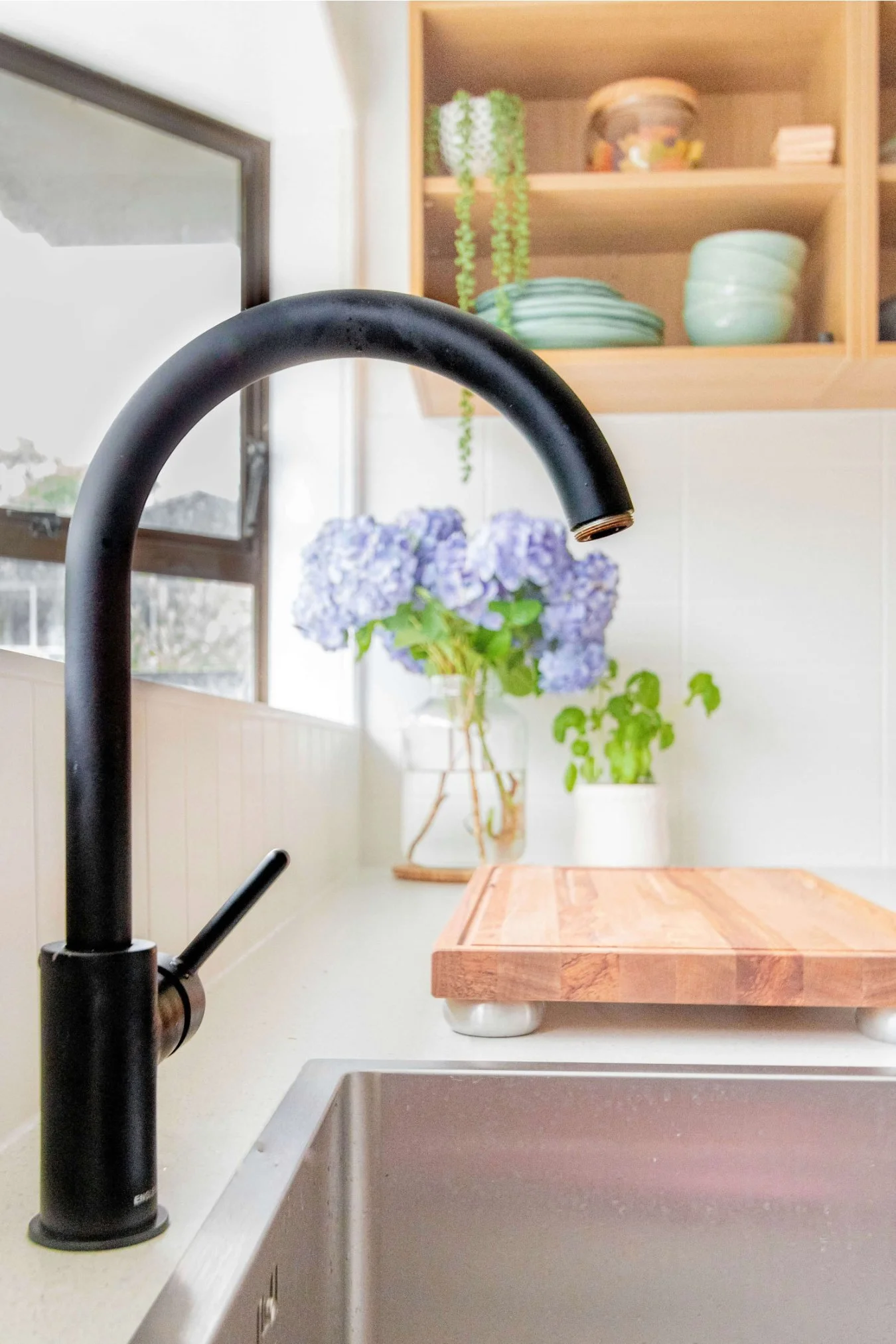 Black kitchen faucet next to window frame with warm wood cabinetry.