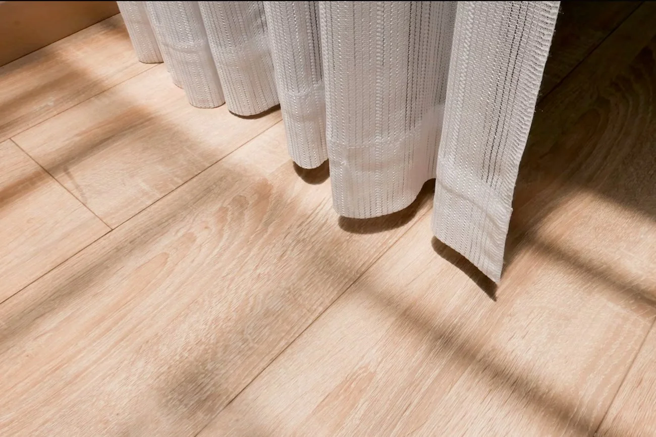 Close-up of a wooden floor with sunlight and shadow filtering through a nearby curtain, highlighting the natural grain and warm texture of the wood.