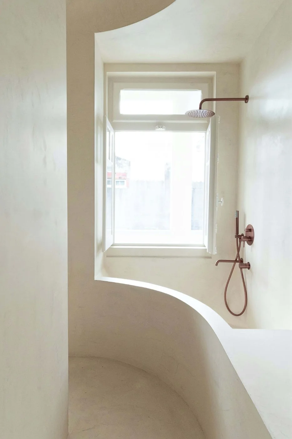 Minimal shower corner with seamless microcement walls and floor beside a large window.