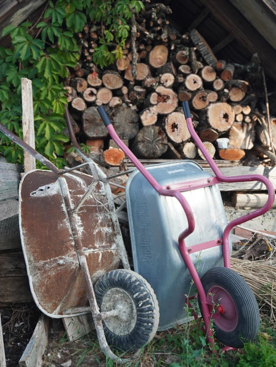 Rustic garden work zone showing heavy-duty wheelbarrows next to a neatly stacked firewood pile and climbing green vines against a natural stone wall.