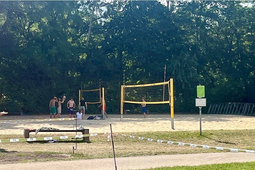 A wide shot of a sand beach volleyball court next to a public swimming lake surrounded by green trees on a sunny summer day.