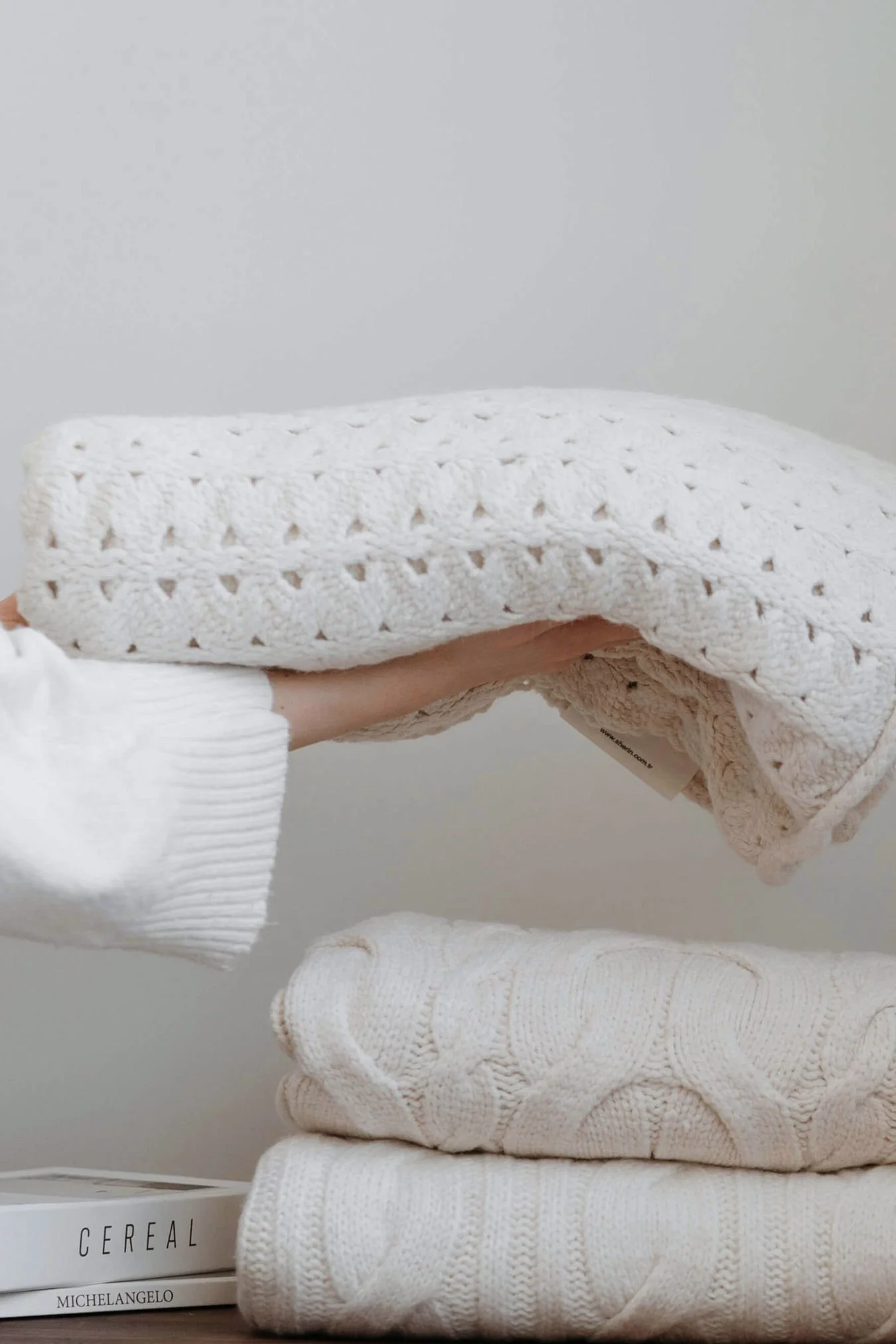 Hands placing a soft folded blanket onto a stack of neutral-toned textiles, highlighting tactile layering and softness.