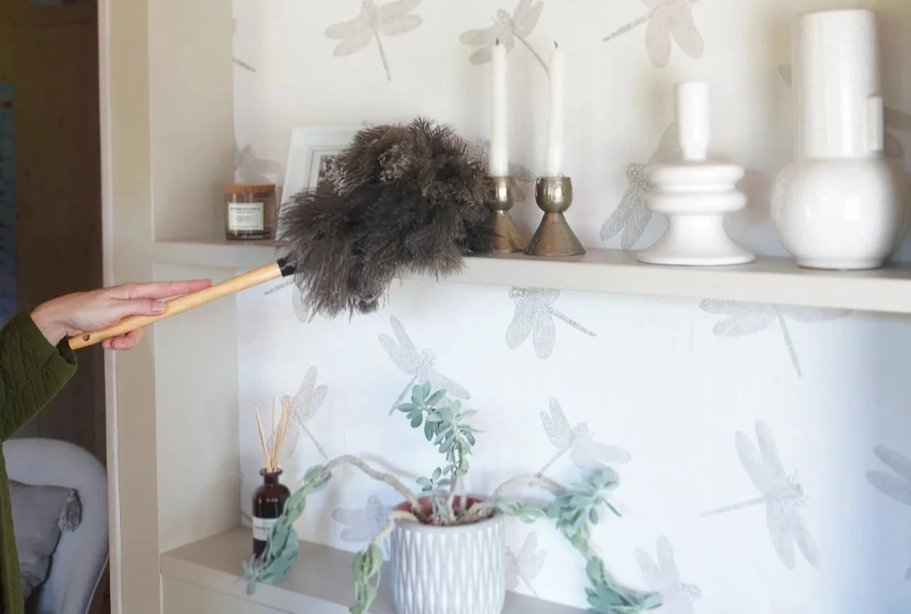 Hands using a feather duster to clean white shelves, representing slow, mindful holiday home preparation and decluttering.