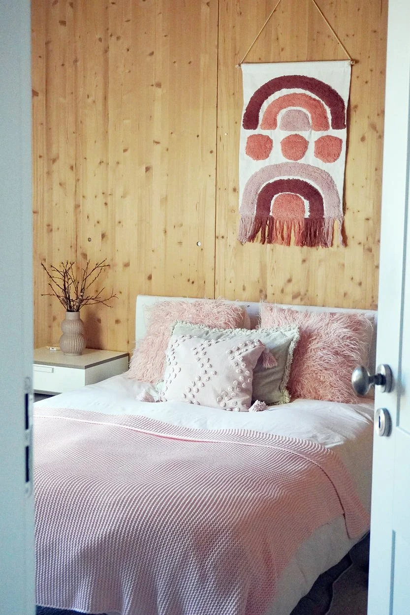 A soft pink boho natural bedroom viewed through an open door, filled with warm daylight